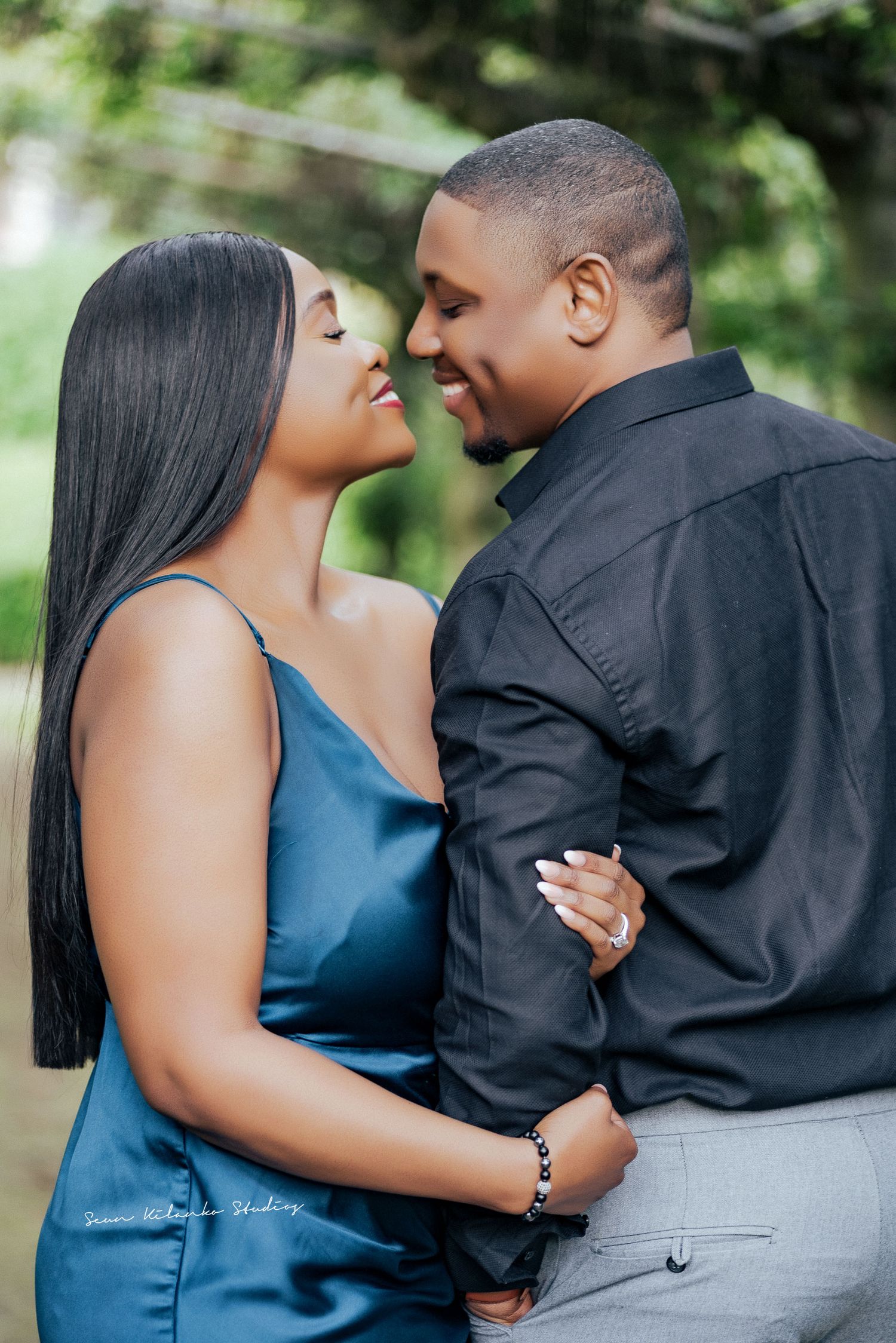 A couple in elegant attire shares a romantic moment outdoors, smiling at each other in a lush green setting.