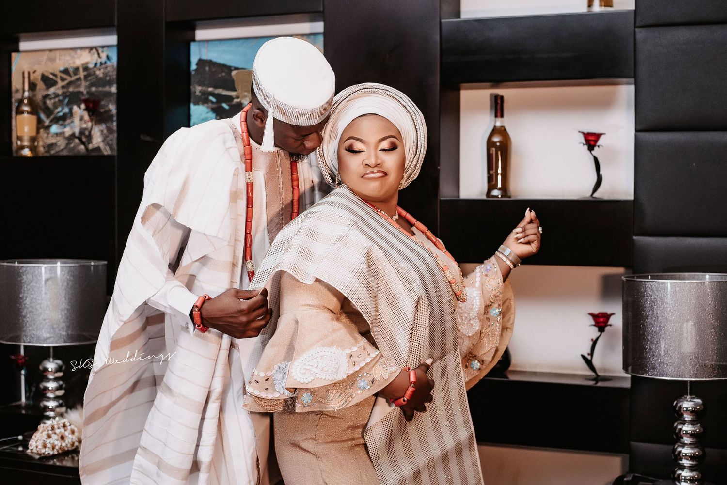A couple in traditional Nigerian cream-colored attire poses together against a modern interior with shelving.