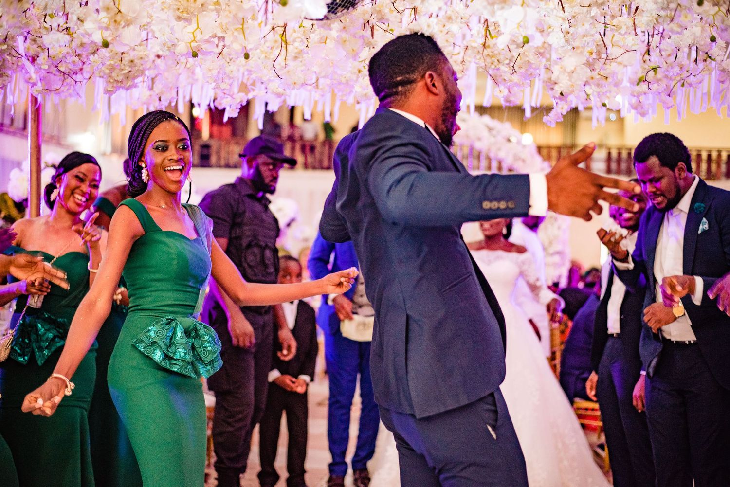 People dancing and celebrating at an elegant party with crystal chandeliers and purple lighting effects above.