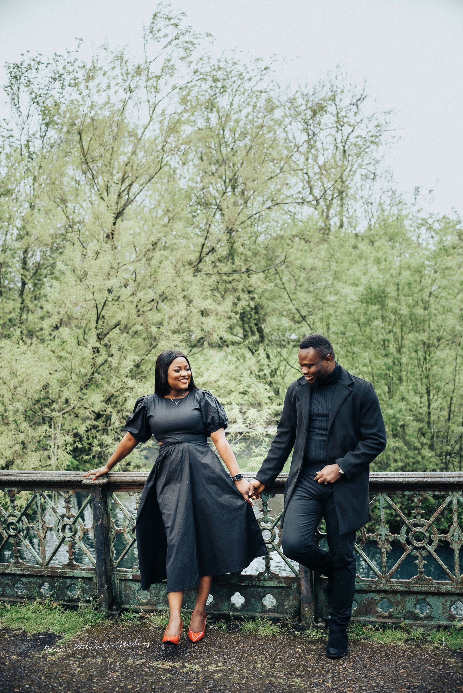 Couple in matching black attire holds hands while posing against a decorative railing in a lush garden setting.