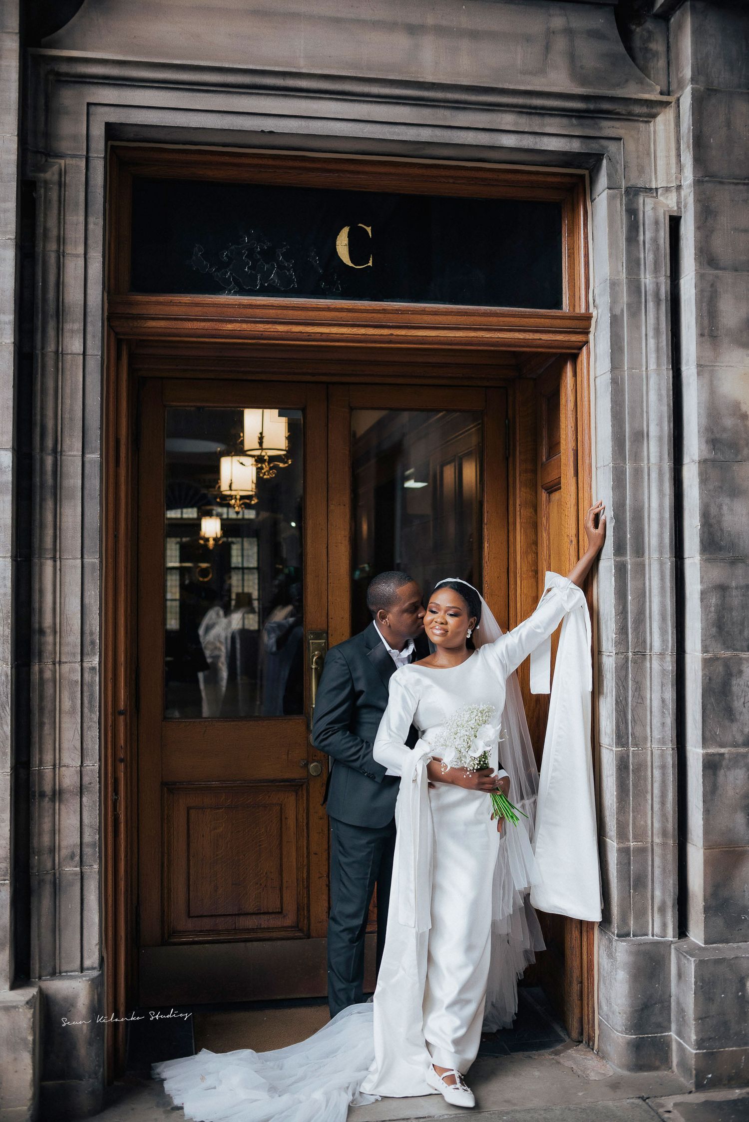 Joyful wedding couple pose in elegant attire at grand entrance doorway.