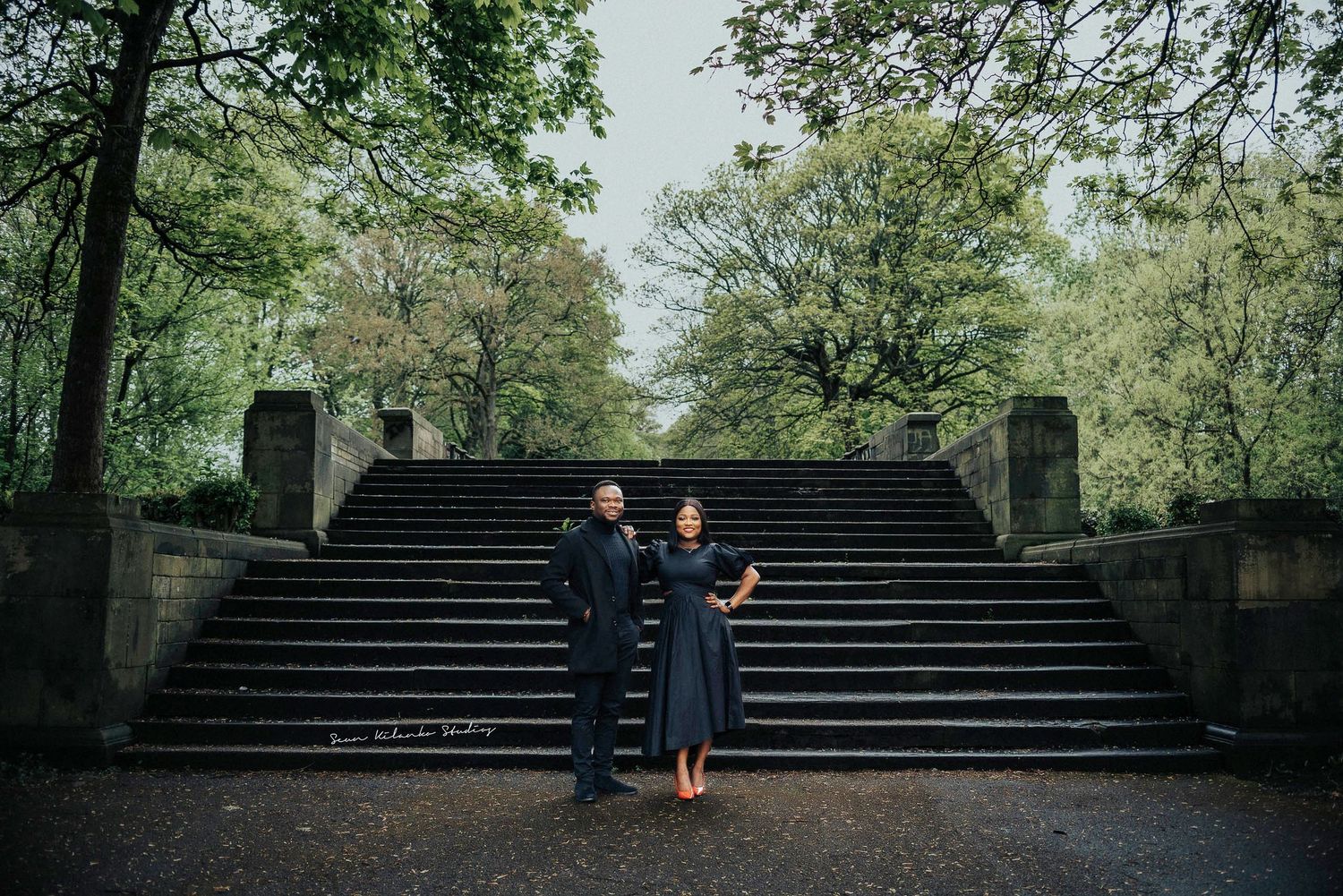 A couple in graduation regalia stands together on stone steps surrounded by green trees in a park setting.