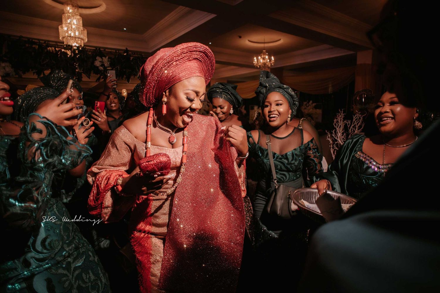 A group of partygoers dance and celebrate in formal attire with chandeliers visible in the background.