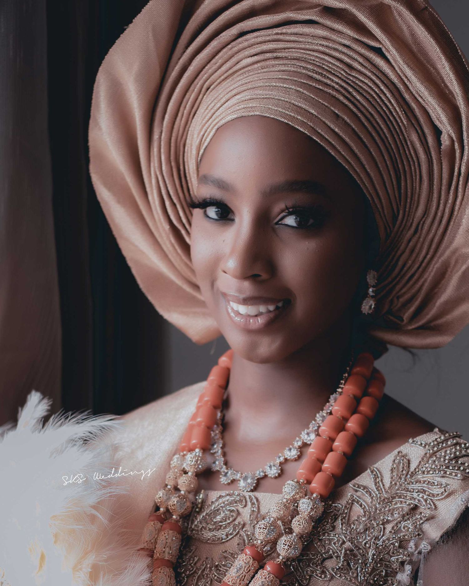 Traditional Nigerian bride wearing a pink gele headwrap, coral beaded necklaces and embroidered dress in elegant portrait.