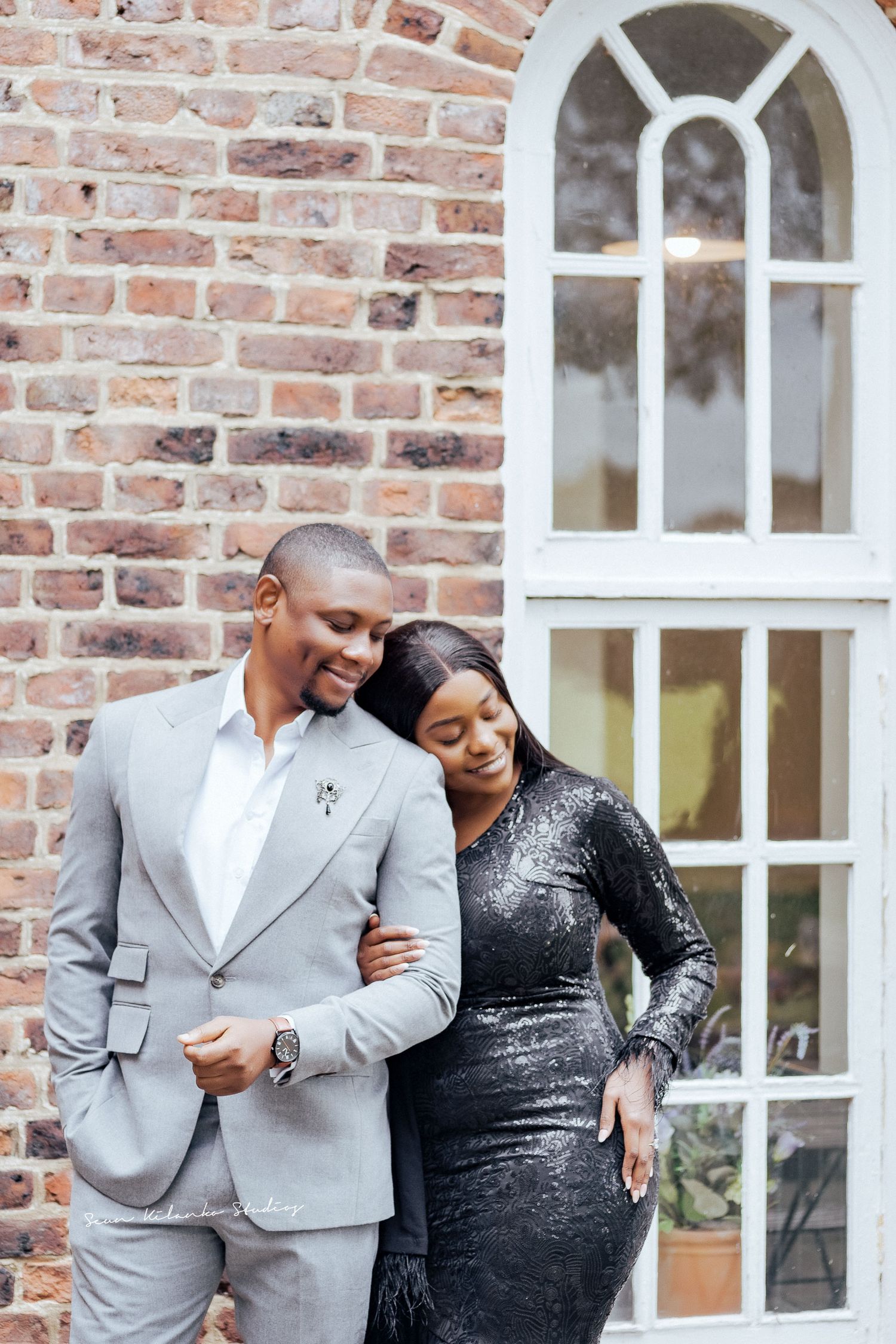 Stylish couple poses by brick wall with arched window, wearing grey suit and black dress.