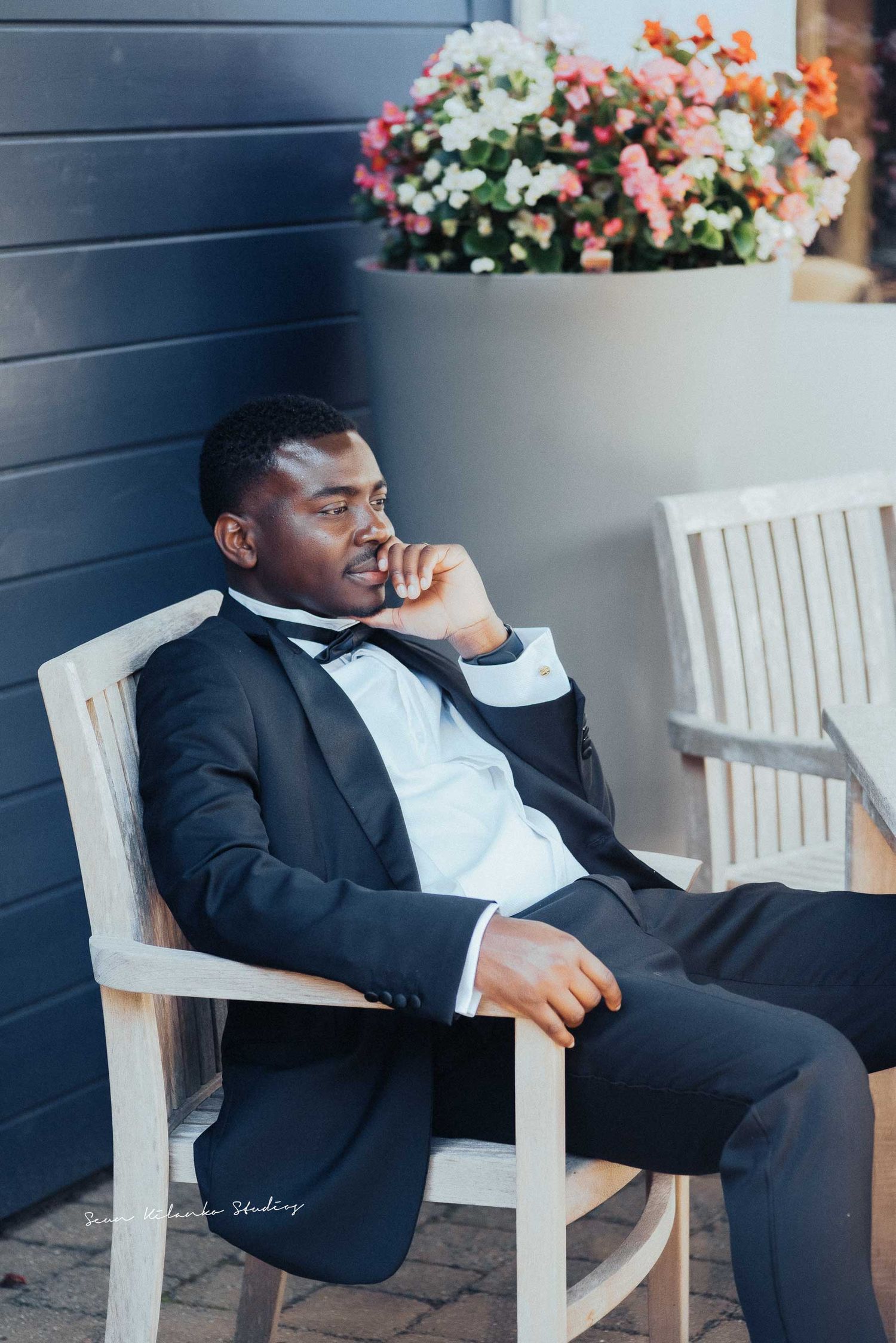 Elegant figure in a tuxedo relaxes thoughtfully in a white chair beside blooming flowers.