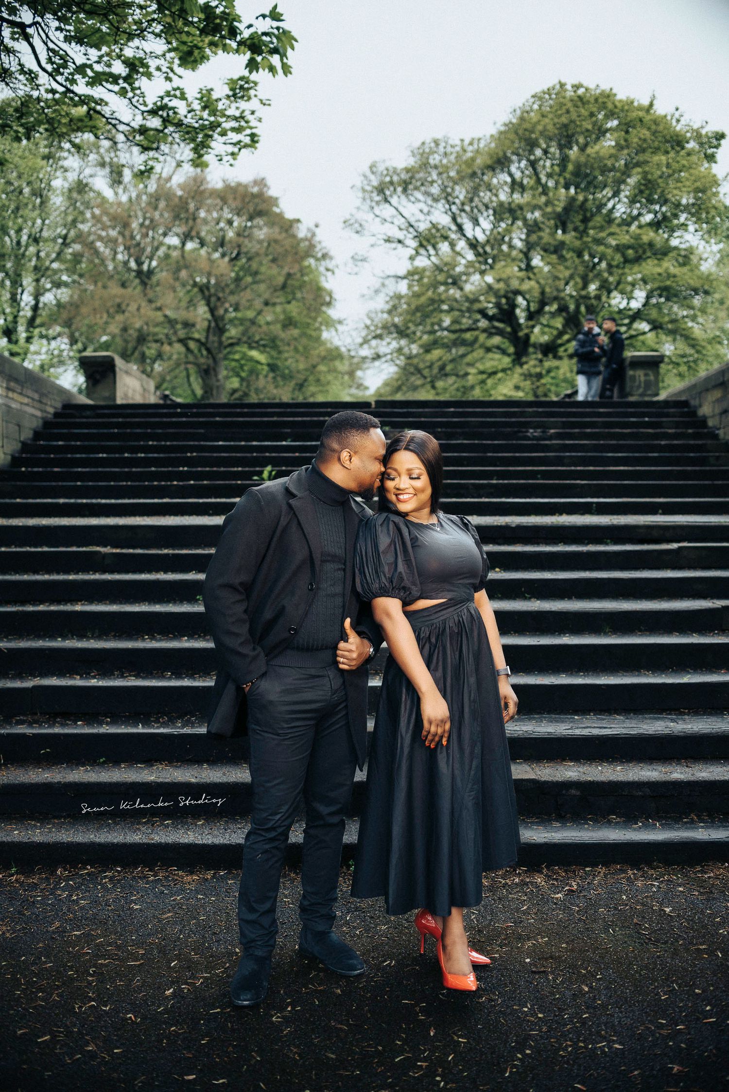 Couple in coordinated black outfits pose intimately on stone steps in a park setting.