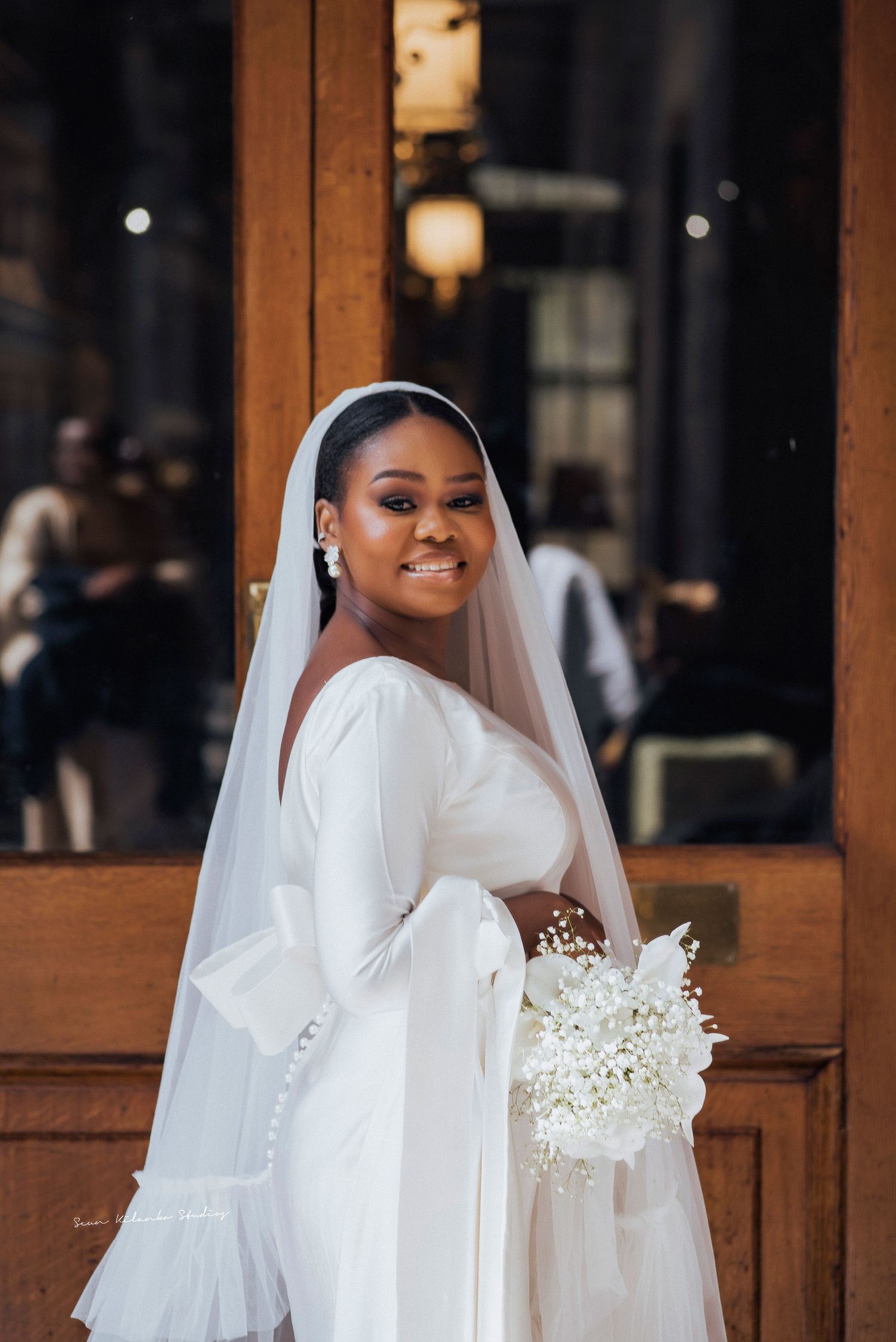 Radiant bride in white long-sleeved gown poses with bouquet near wooden doorframe.