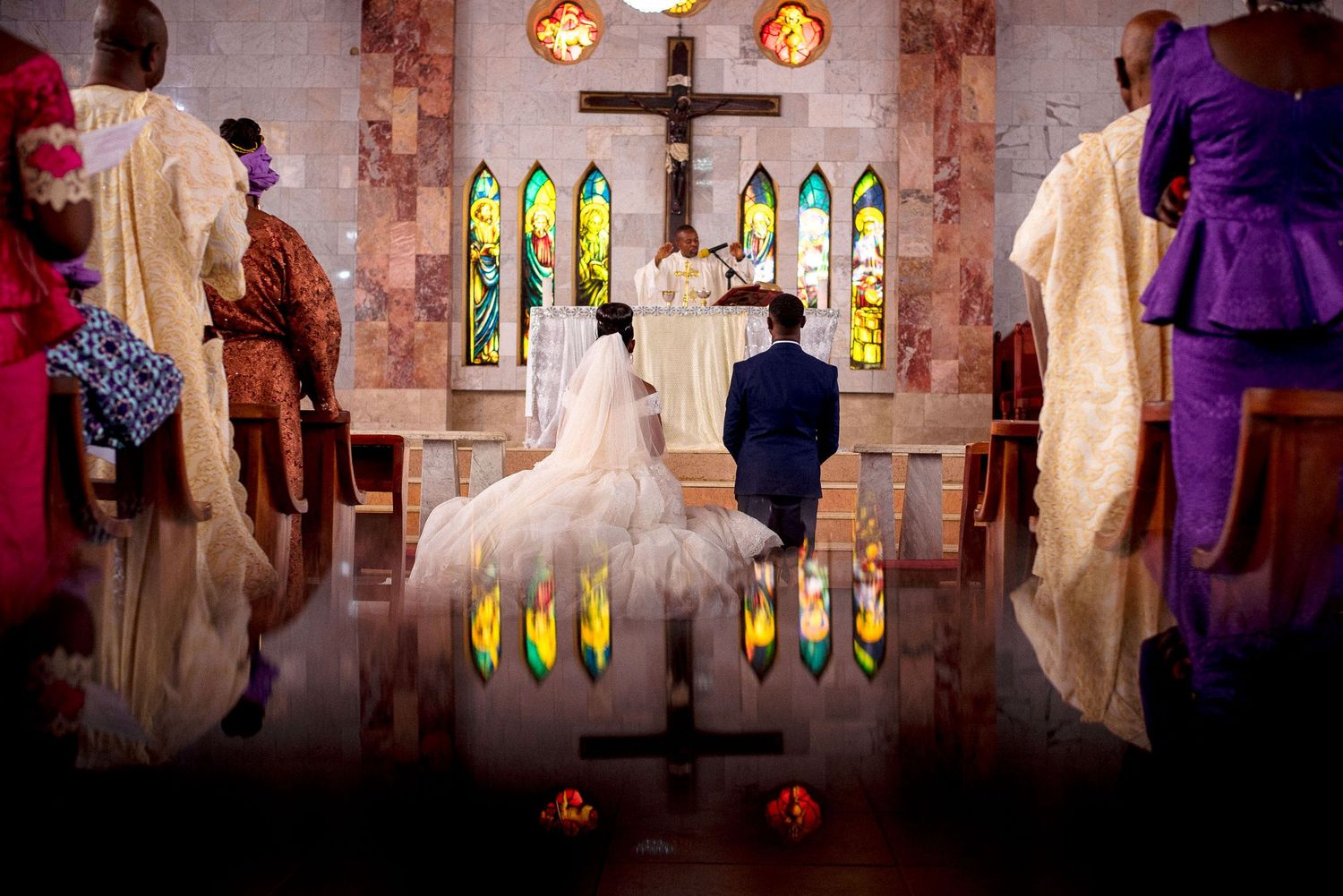 A wedding ceremony with a stained glass backdrop reflects beautifully in mirrored flooring during an intimate church service.