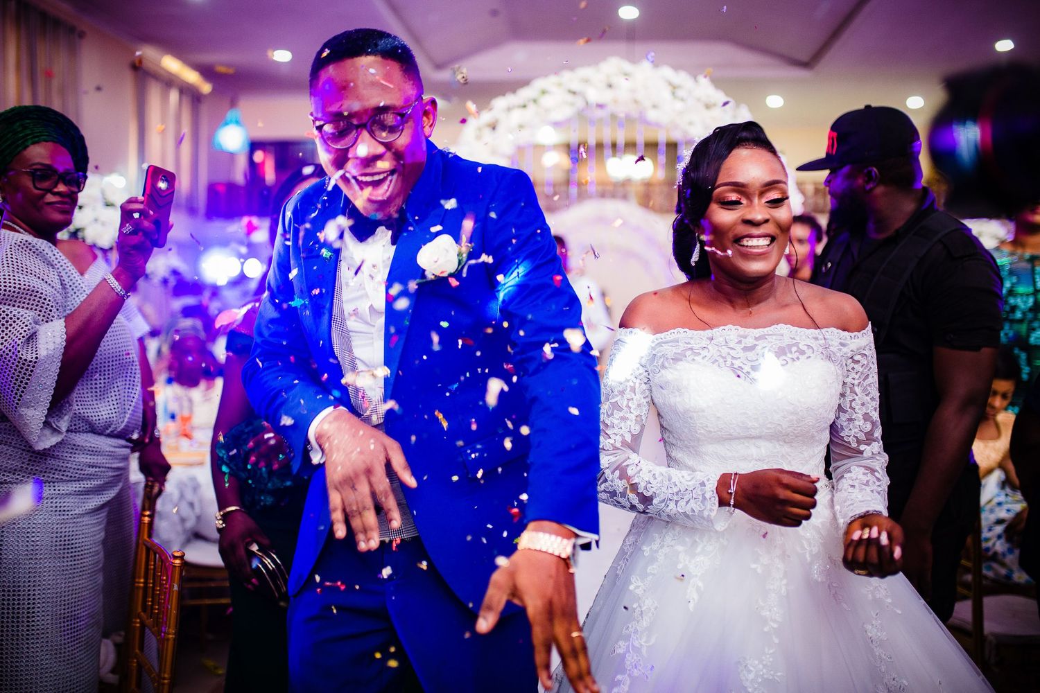 Newlywed couple dances joyfully at their wedding reception as guests cheer and blue lights illuminate the venue.