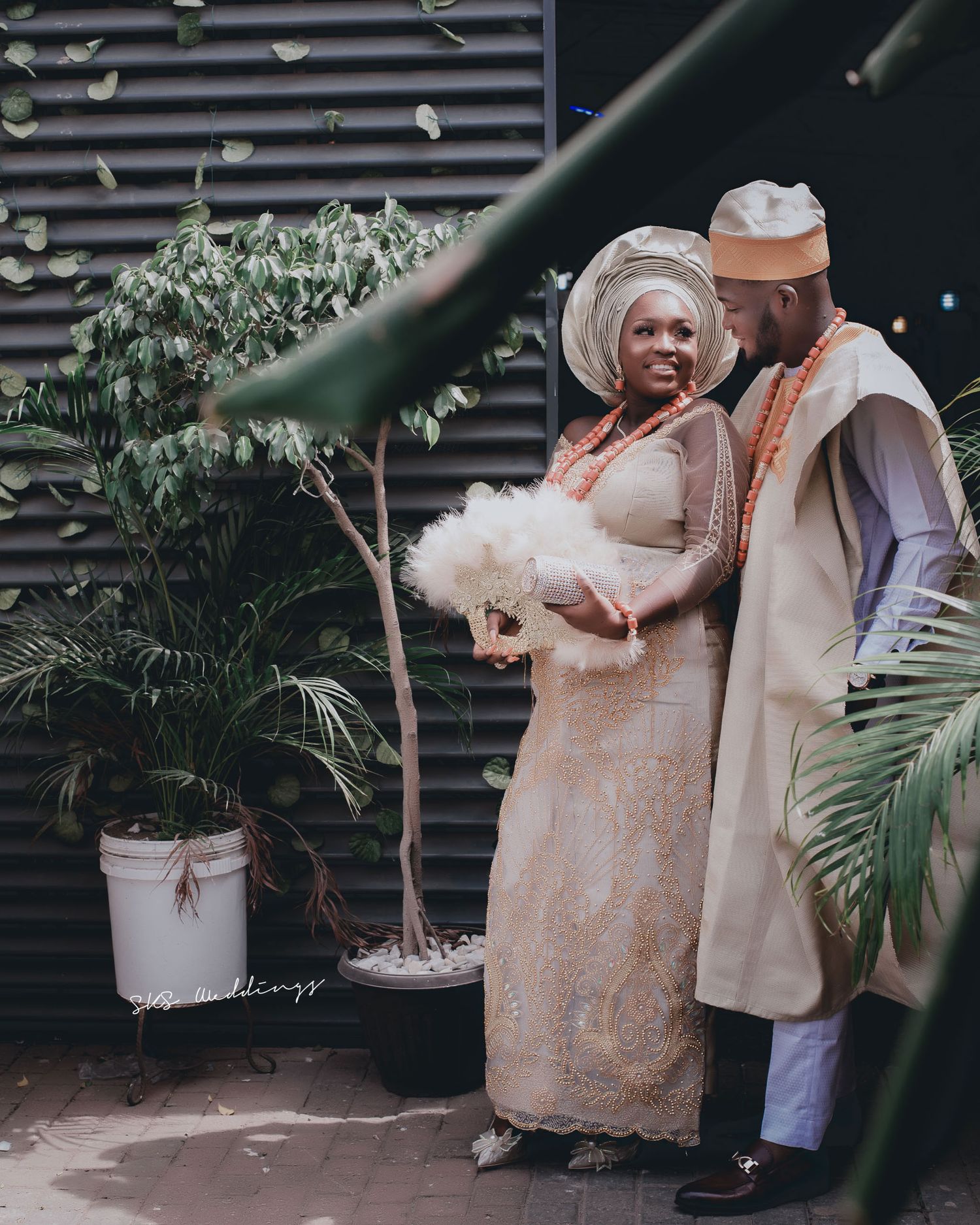 Traditional Nigerian couple in elegant cream and orange attire pose together near potted palm plants against dark wall.