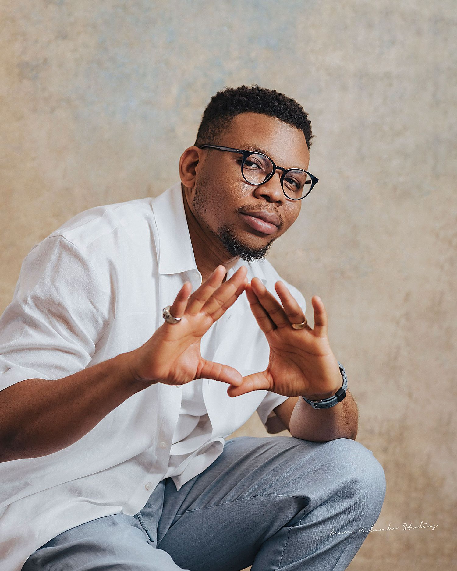 Portrait of a person in glasses and white shirt making expressive hand gestures against a neutral backdrop.