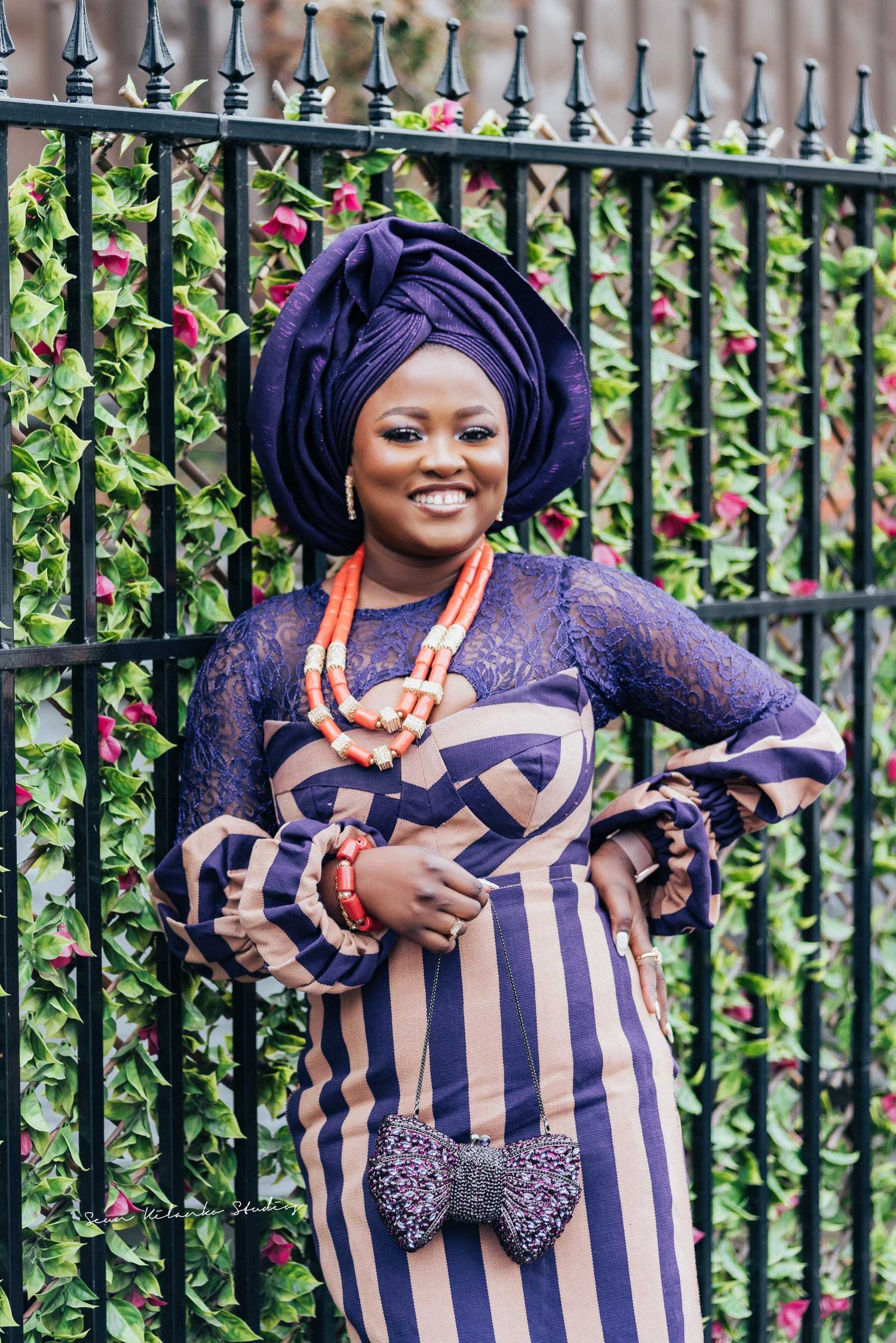 Traditional Nigerian outfit with purple gele and striped ankara dress posed by decorative iron fence.