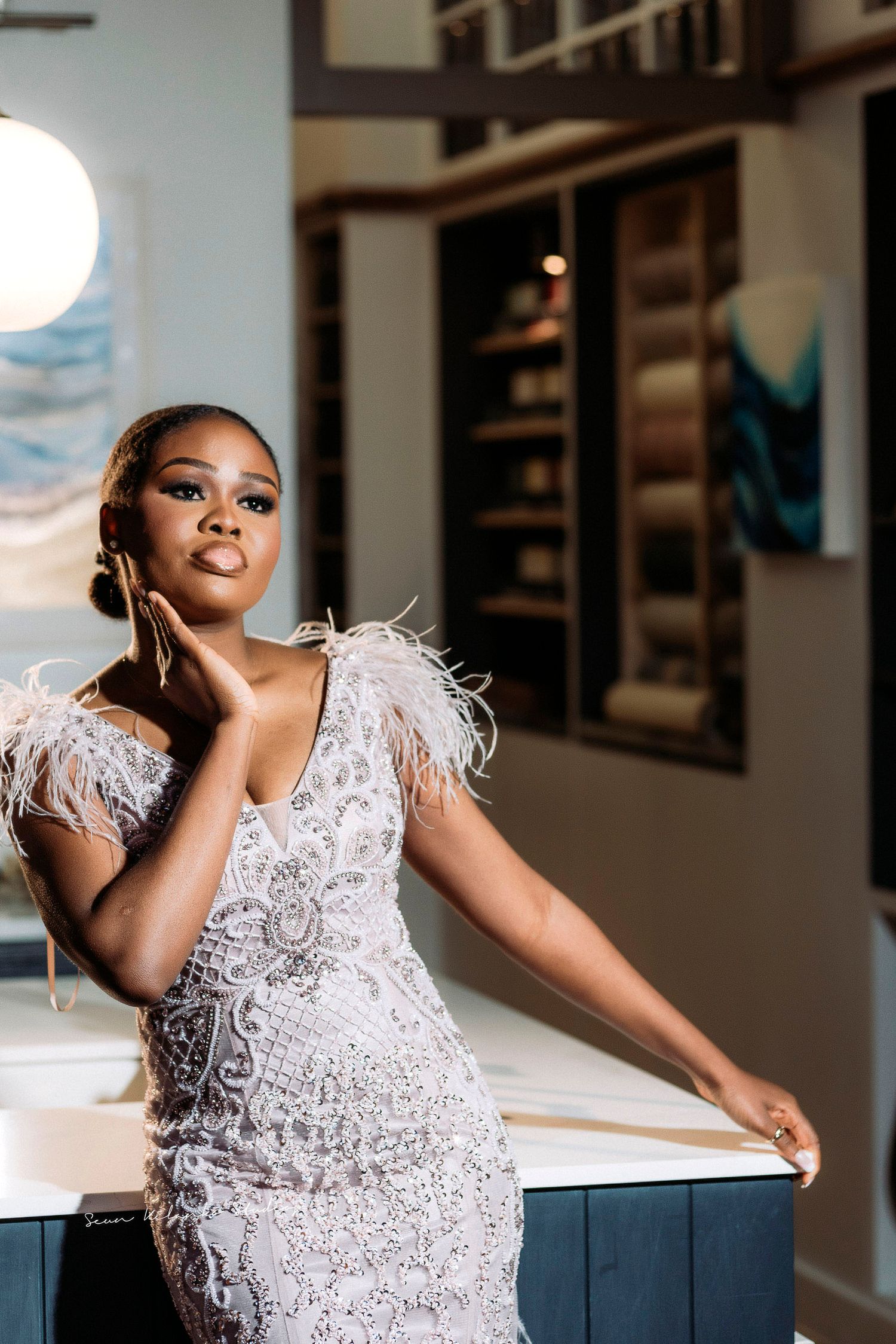 Glamorous bride poses in feather-detailed lace dress against modern interior setting.