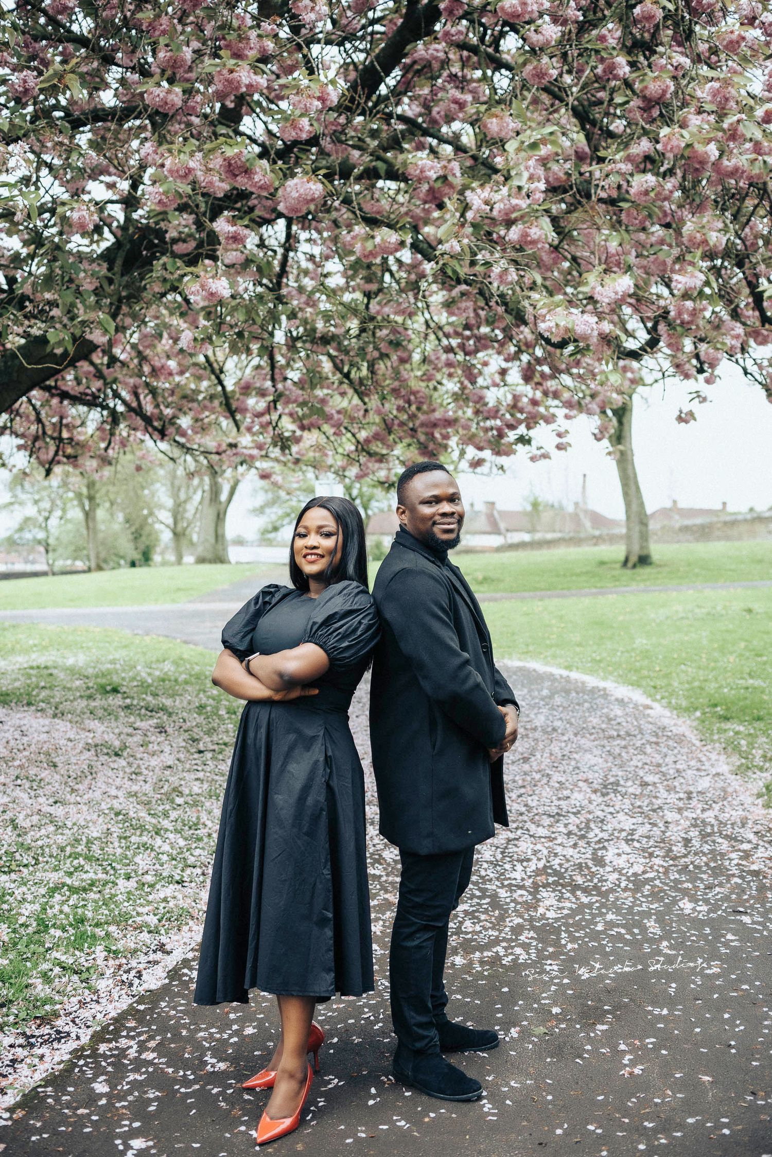 Stylishly dressed couple poses back-to-back under pink cherry blossoms.