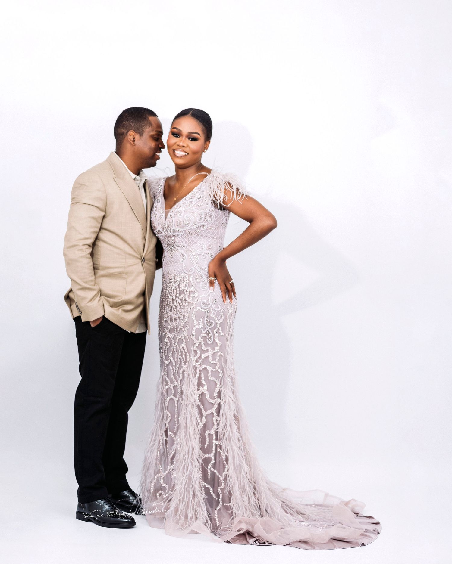 Elegant couple poses in formal attire with bride wearing flowing lace gown against white backdrop.