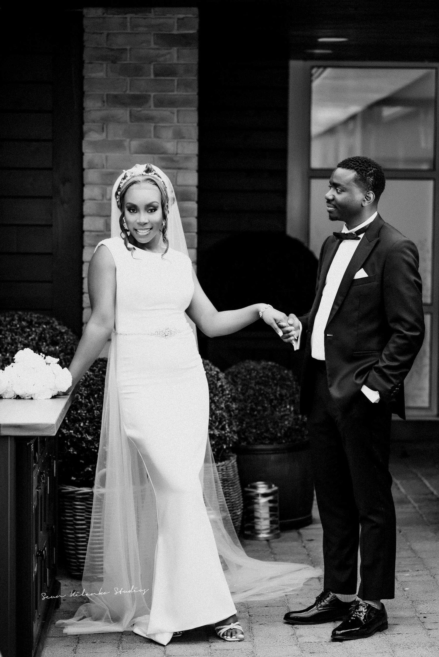 Black and white photo of newlyweds in formal attire sharing a playful moment against a brick wall.