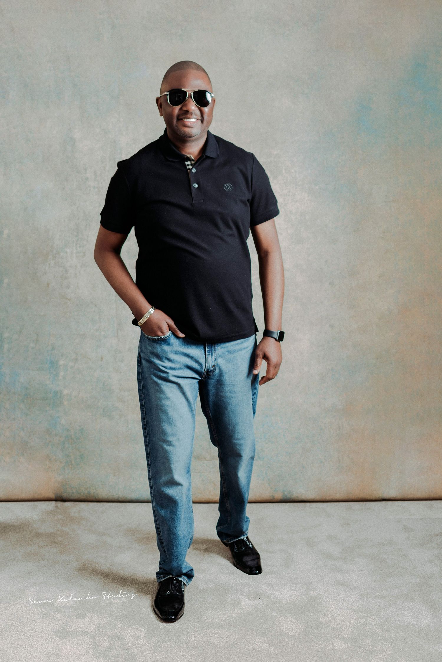 Person in black polo shirt and light jeans poses confidently with sunglasses in studio setting.