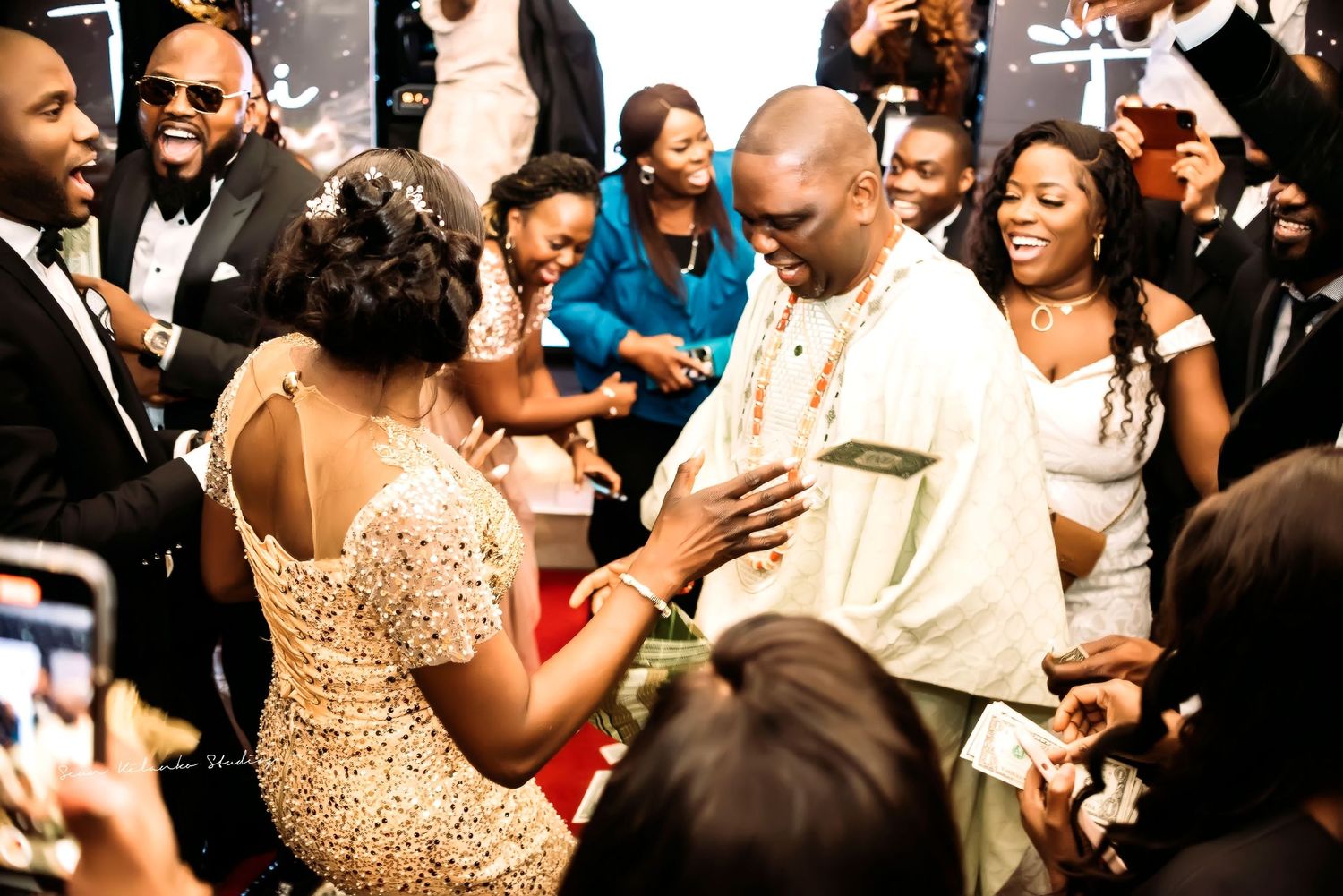 Guests celebrate and dance together at a lively wedding reception with people mingling and smiling in the background.