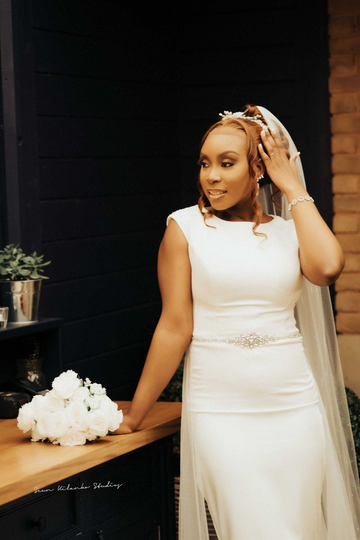 A bride in a sleek white wedding dress with a beaded belt poses elegantly near a wooden shelf.