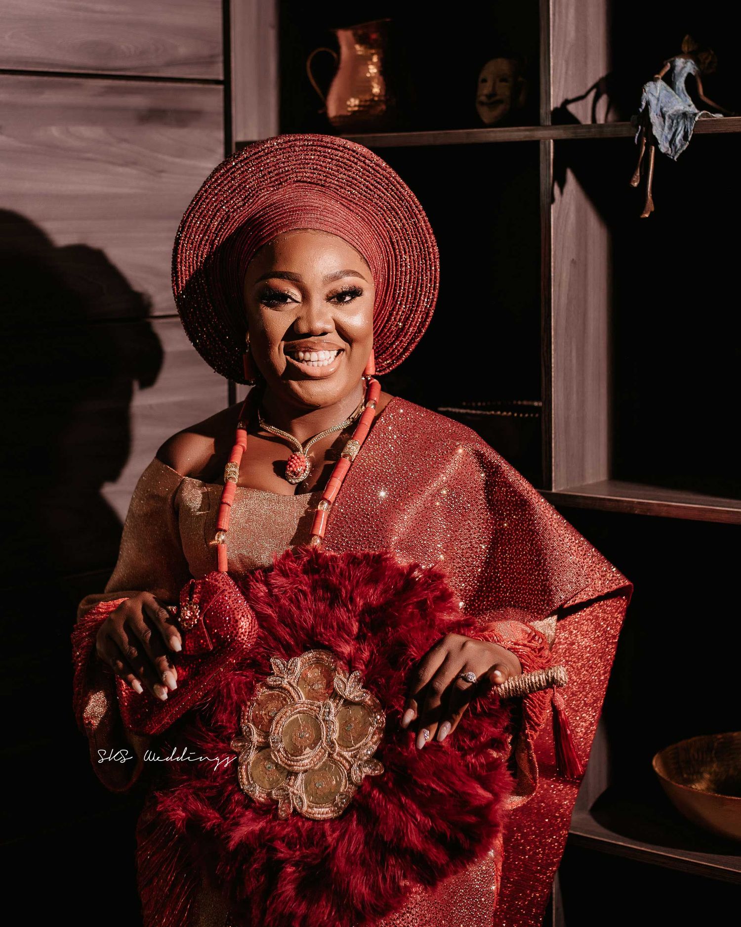 A person in a vibrant red traditional Nigerian gele and dress holds a bouquet of deep red flowers in a warm-toned setting.