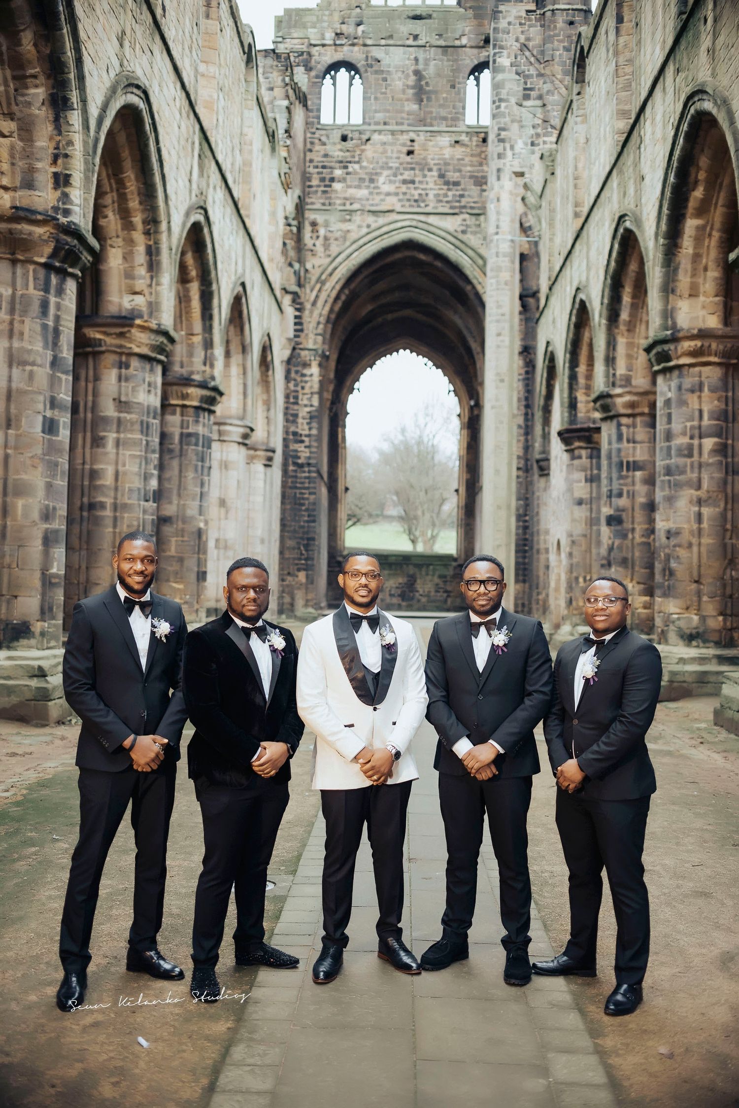 Groomsmen in formal black tuxedos pose together in front of a gothic stone cathedral ruins.