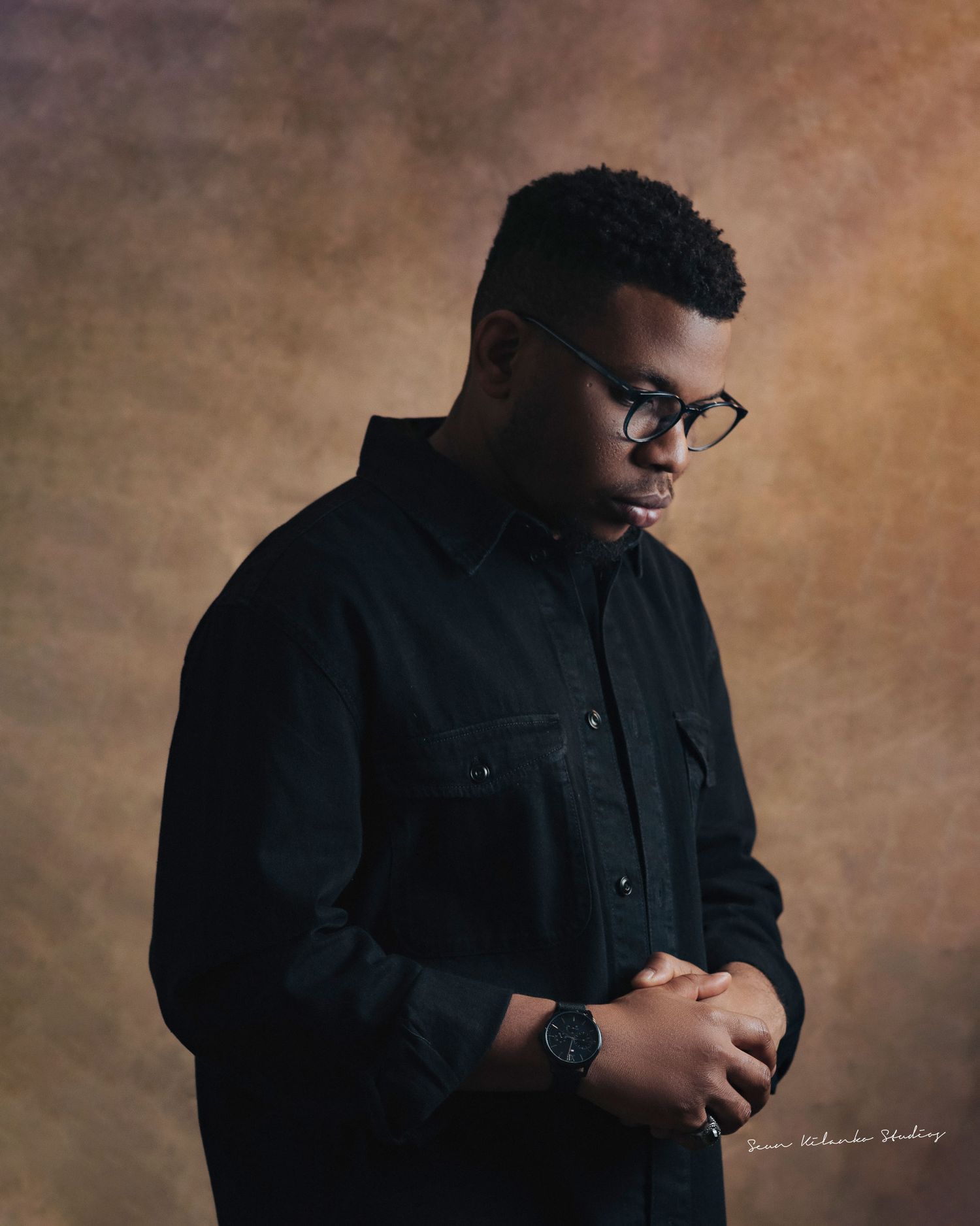 Professional studio portrait of someone in a black button-up shirt and glasses against a brown backdrop.