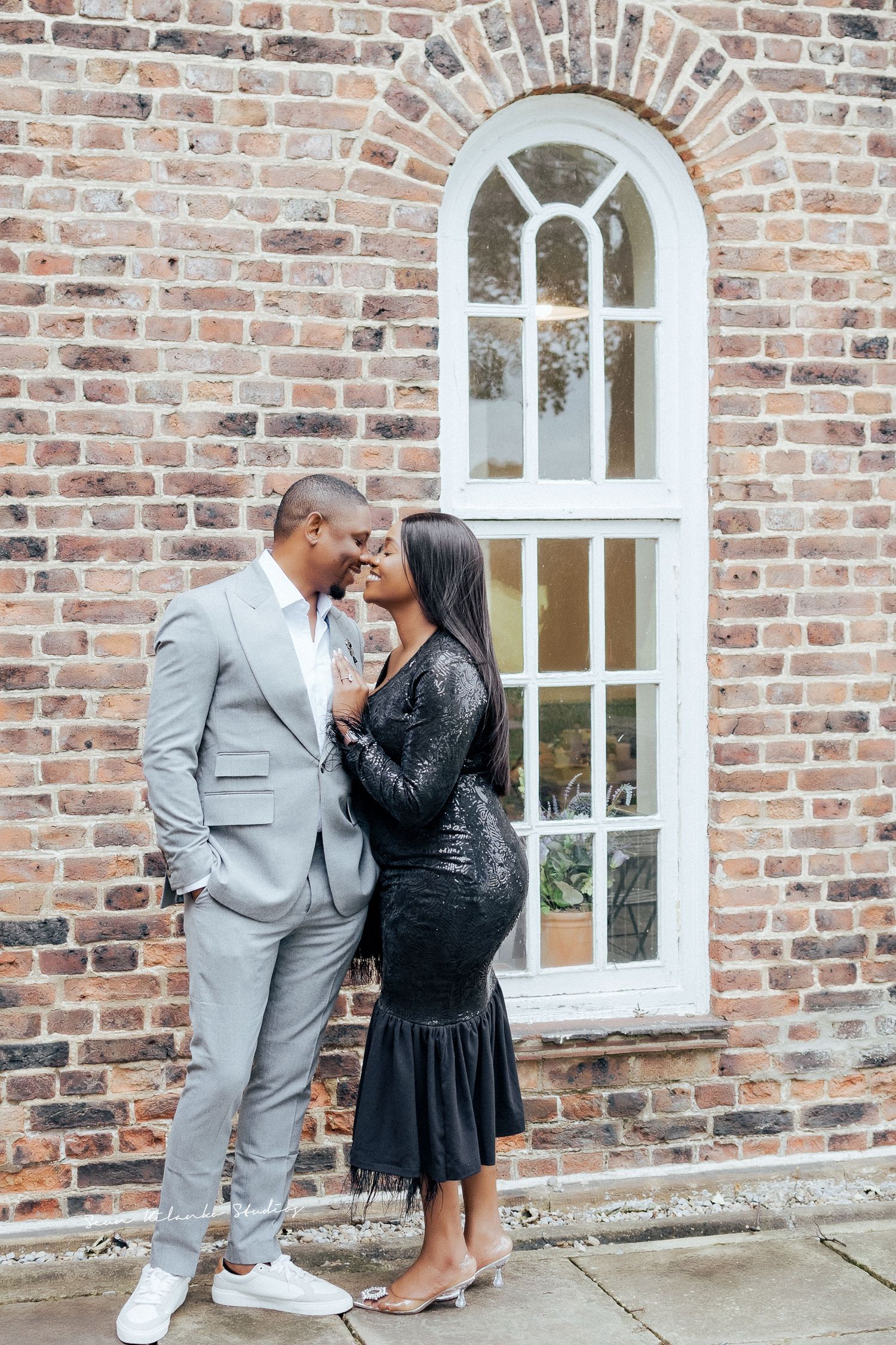 Couple in formal attire poses intimately against a brick wall with an arched window.