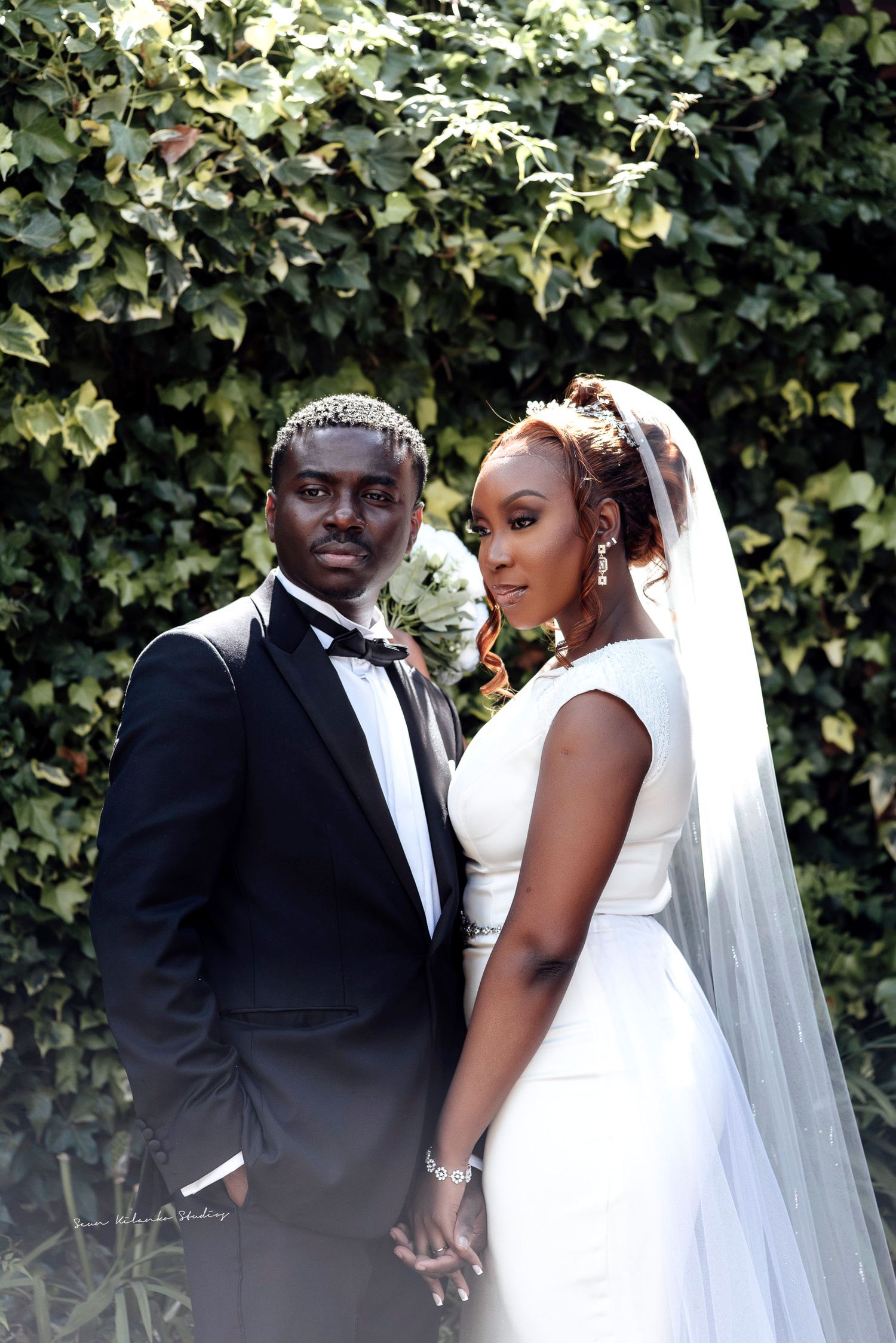 A bride and groom pose elegantly in front of a lush ivy wall, she in a white gown with veil, he in a black tuxedo.