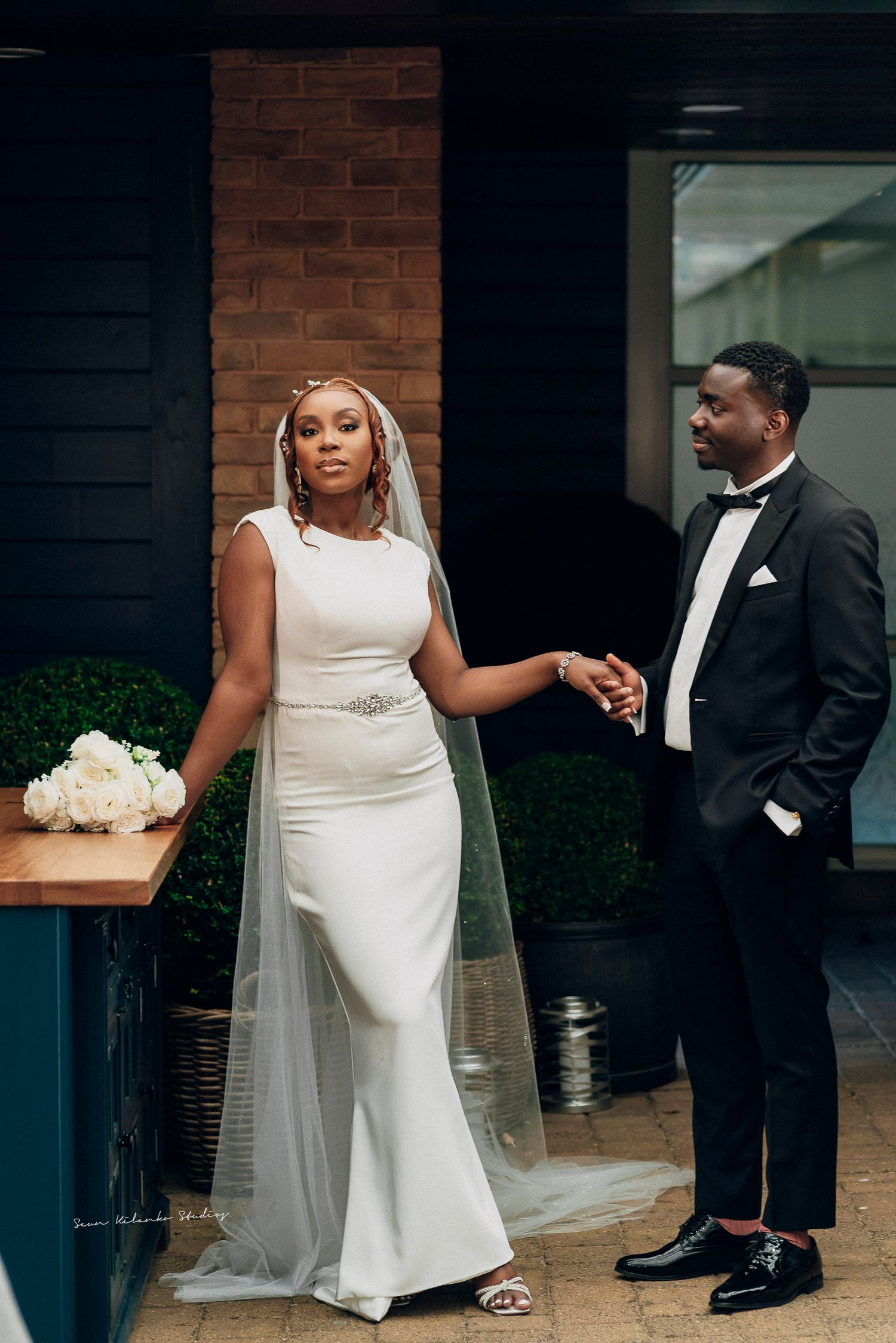 A wedding couple in white dress and black suit hold hands in an intimate moment against a brick wall backdrop.