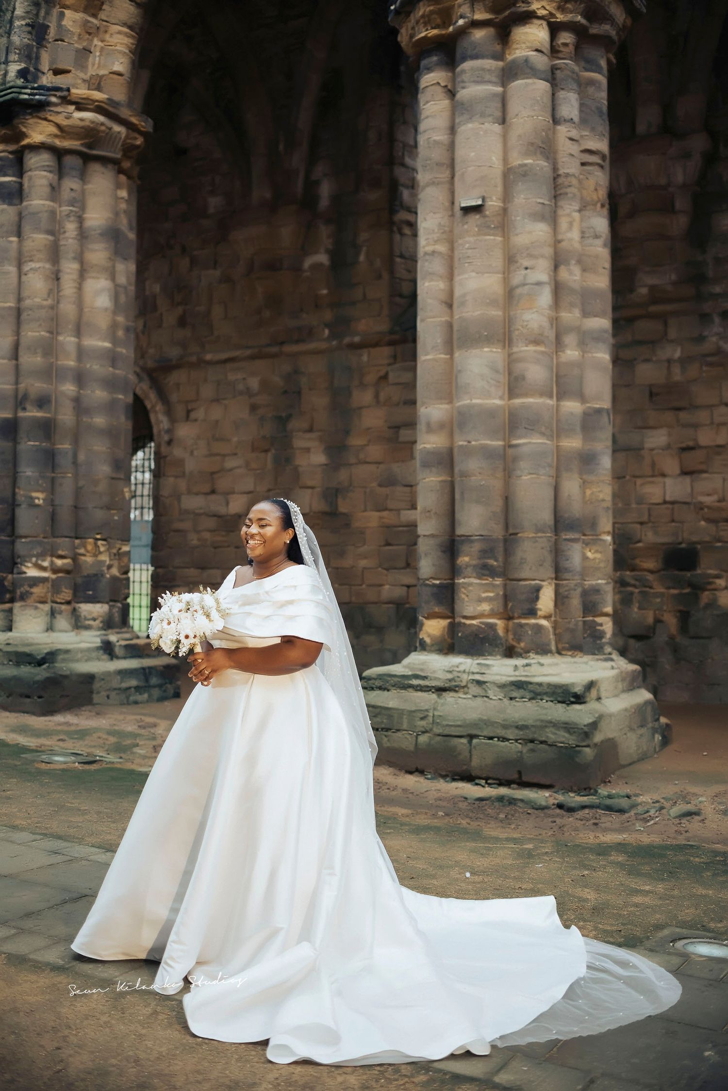 A radiant bride holds a bouquet while standing beneath historic stone cathedral columns.