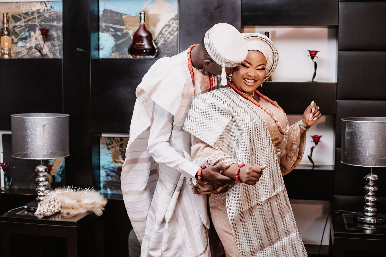 Two people in white garments embrace in celebration with decorative table lamps visible in background.