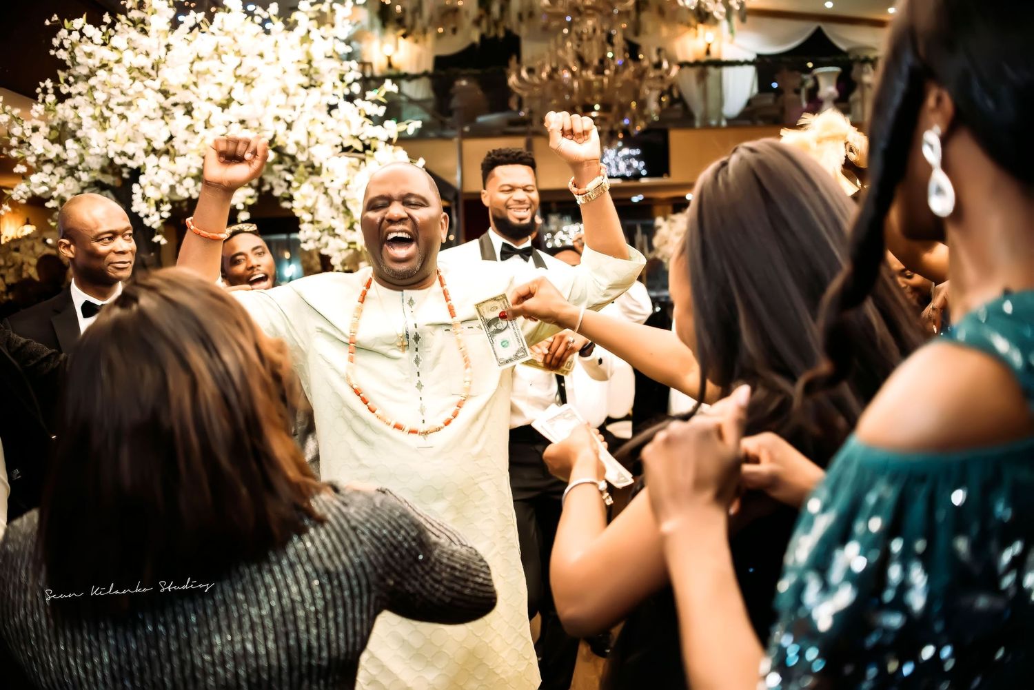 Wedding guests dance and celebrate joyfully under glowing lights at an indoor reception party.
