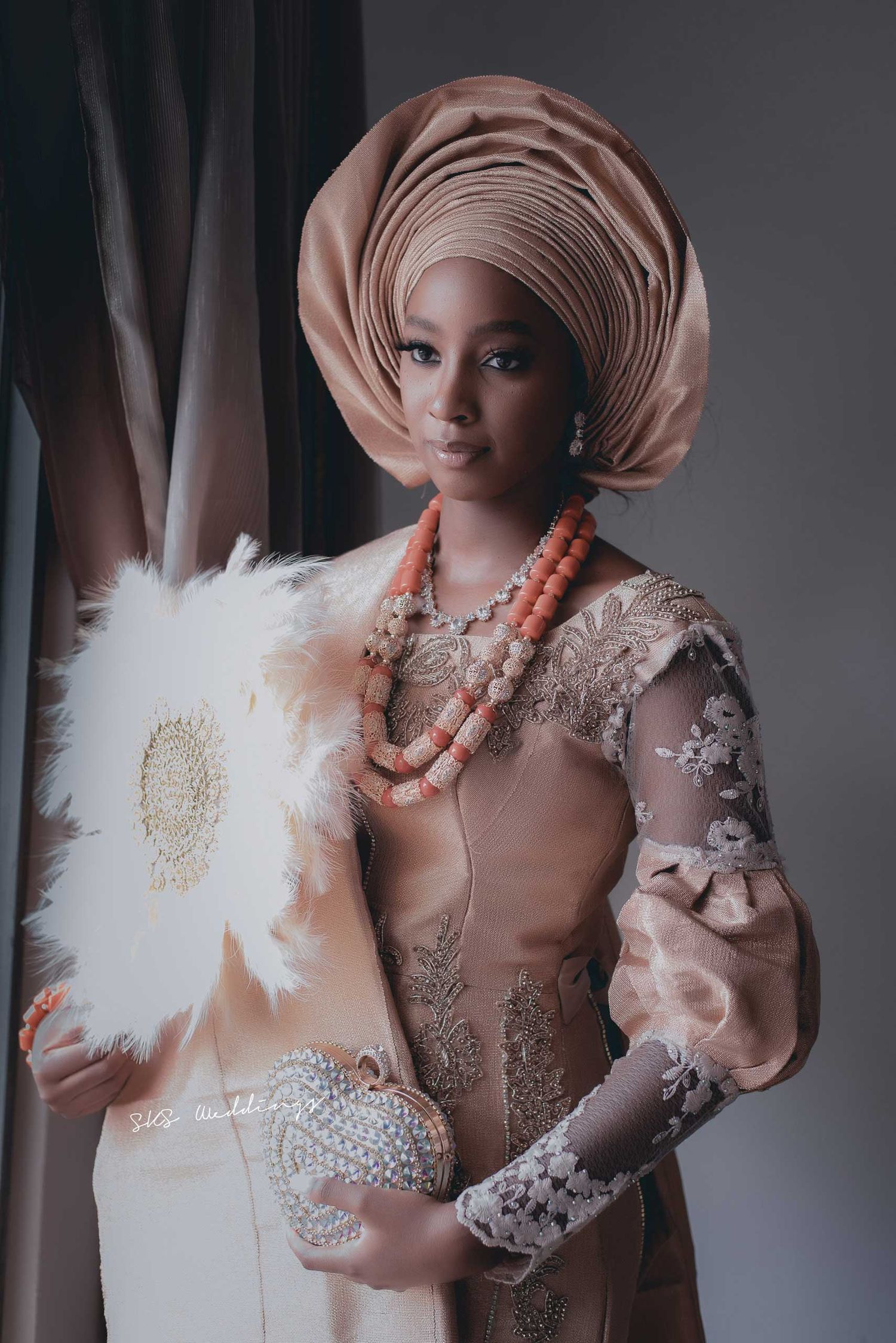 Traditional Nigerian bride wearing beige gele headwrap, coral beads and lace dress holding ceremonial hand fan.