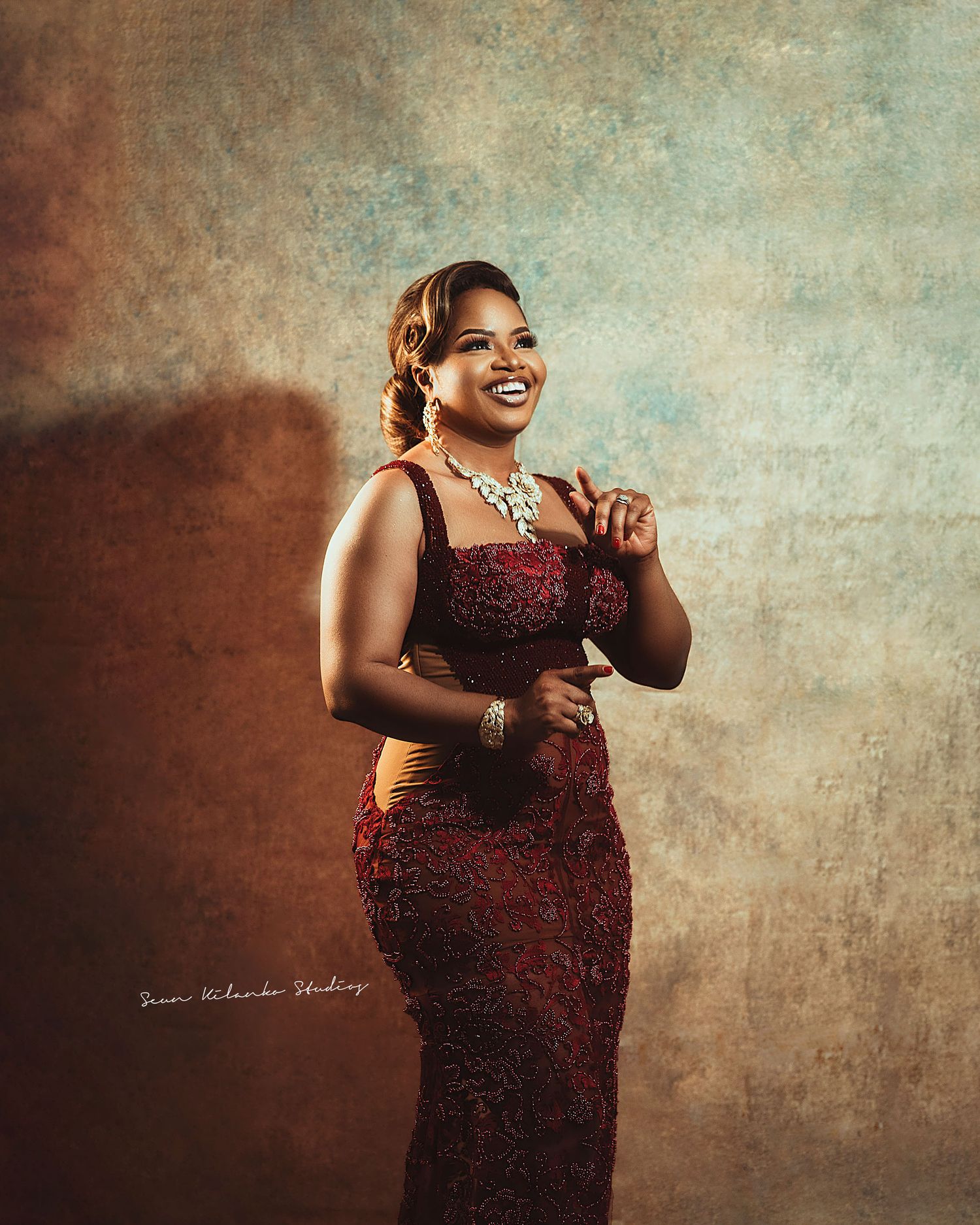 Joyful portrait in a form-fitting burgundy gown with crystal necklace against vintage backdrop.