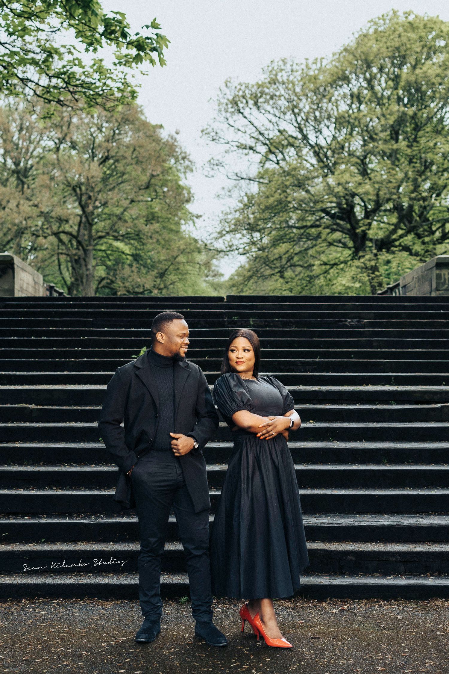 Elegant couple in black outfits poses confidently on grand stone steps with trees in background.