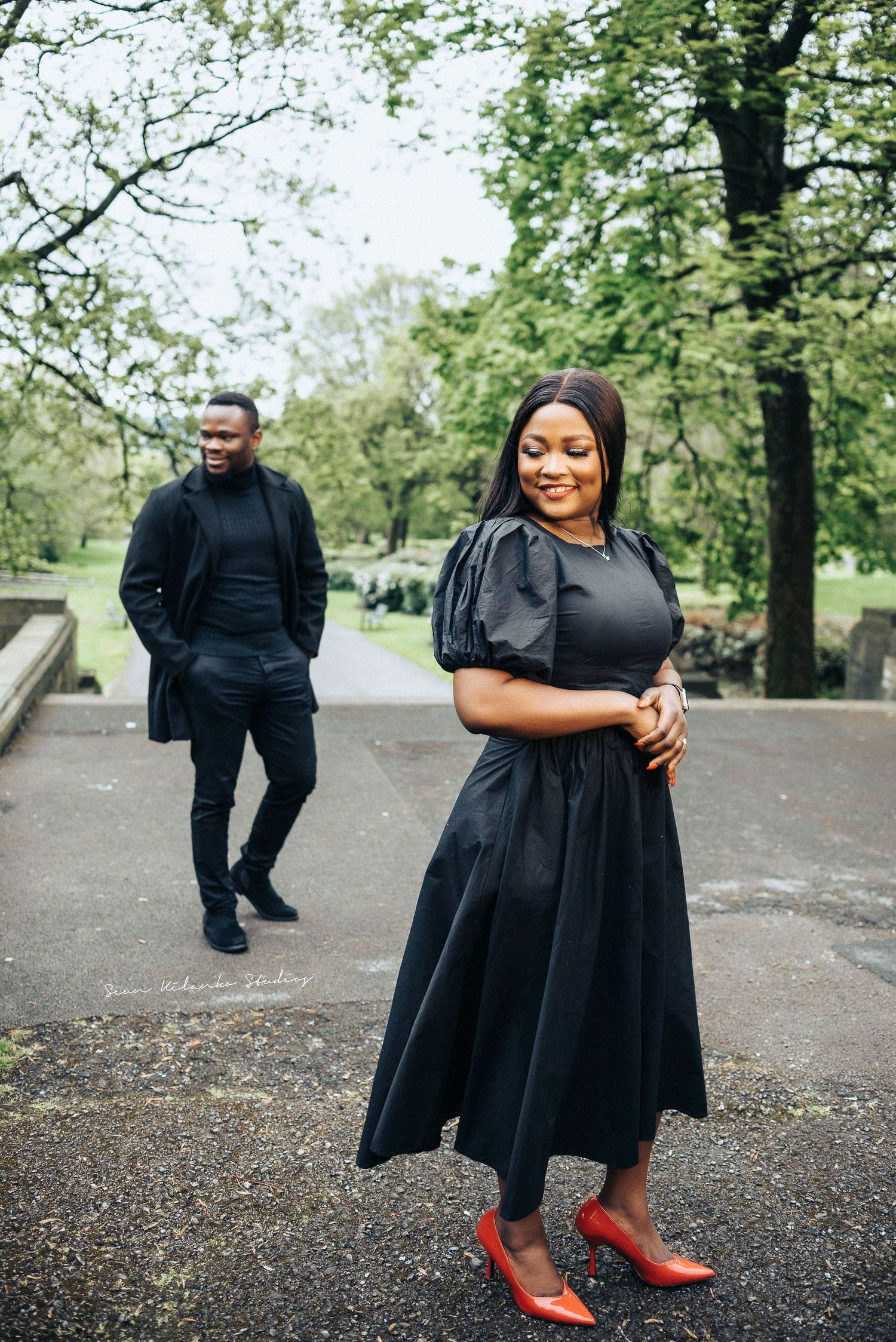 A stylish couple in coordinated black outfits poses in a park setting, with the woman wearing bright red shoes.