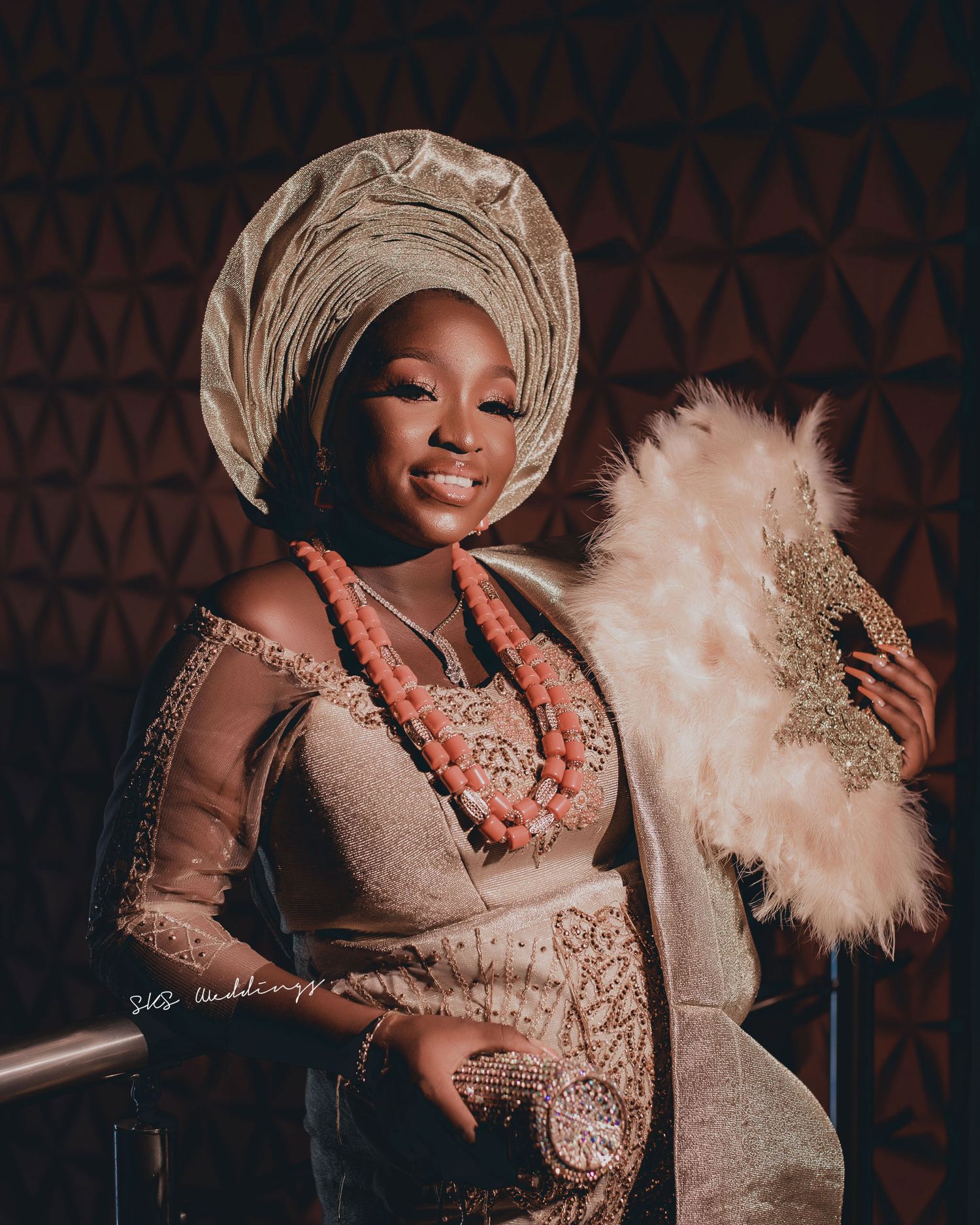 Traditional Nigerian attire featuring gold lace gele headwrap and beaded coral necklaces with a decorative feather fan.