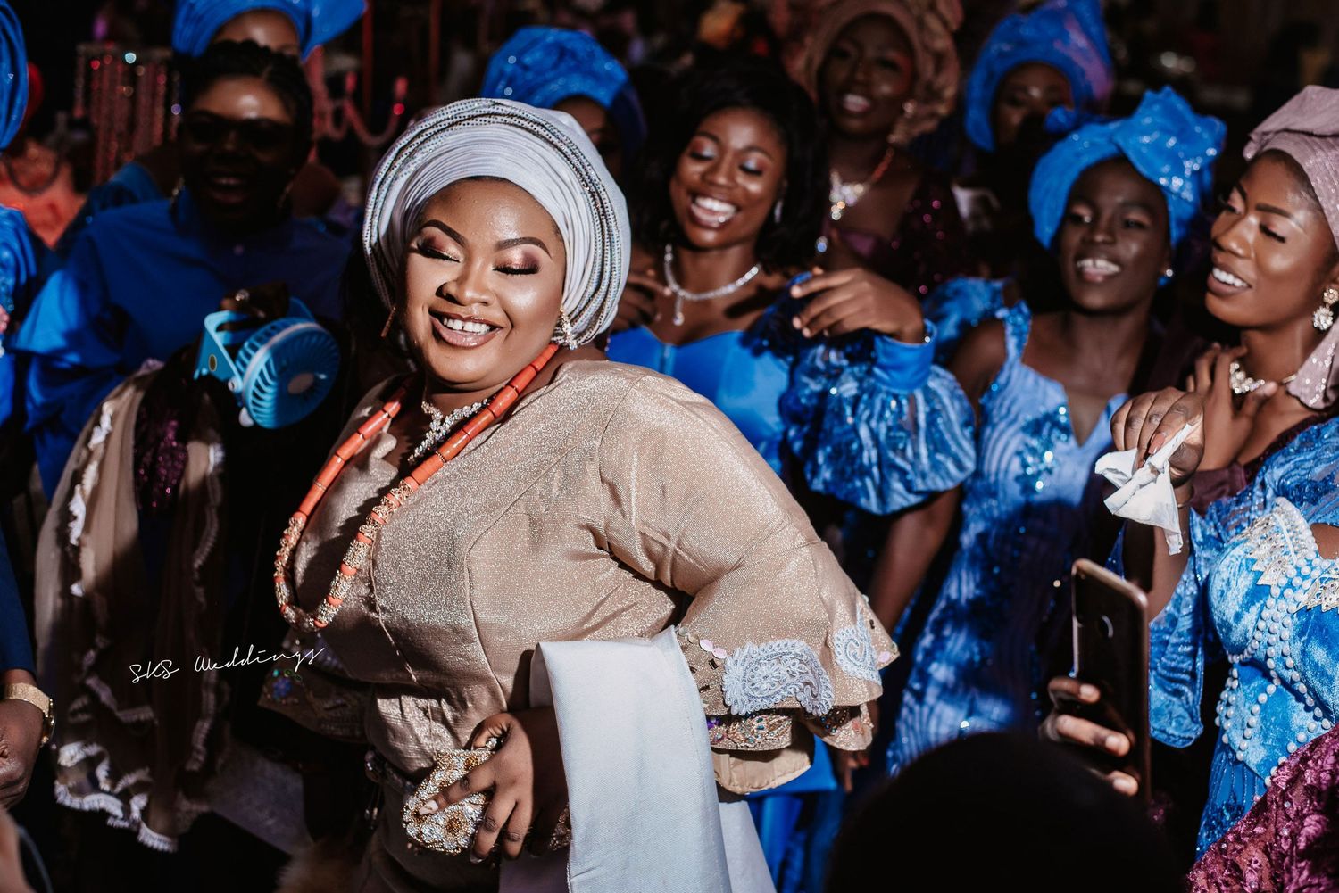 A joyful celebration scene with people in traditional West African attire including blue dresses and beige headwraps.