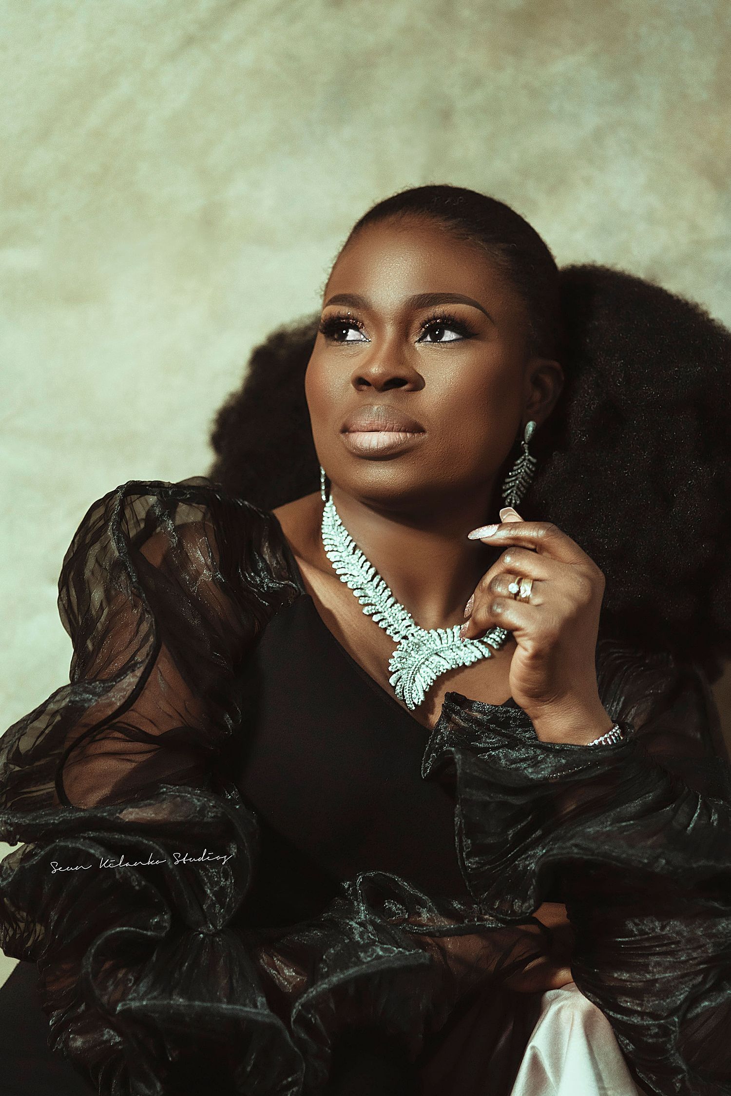 Studio portrait featuring elegant black dress, turquoise statement necklace and dramatic makeup against textured background.