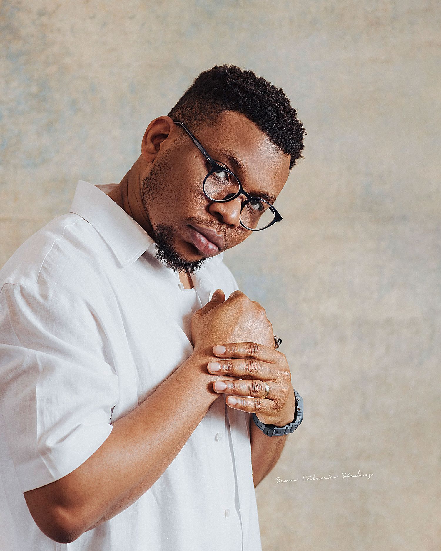 Emotive portrait of a person in glasses and white shirt with clasped hands in prayer-like pose.