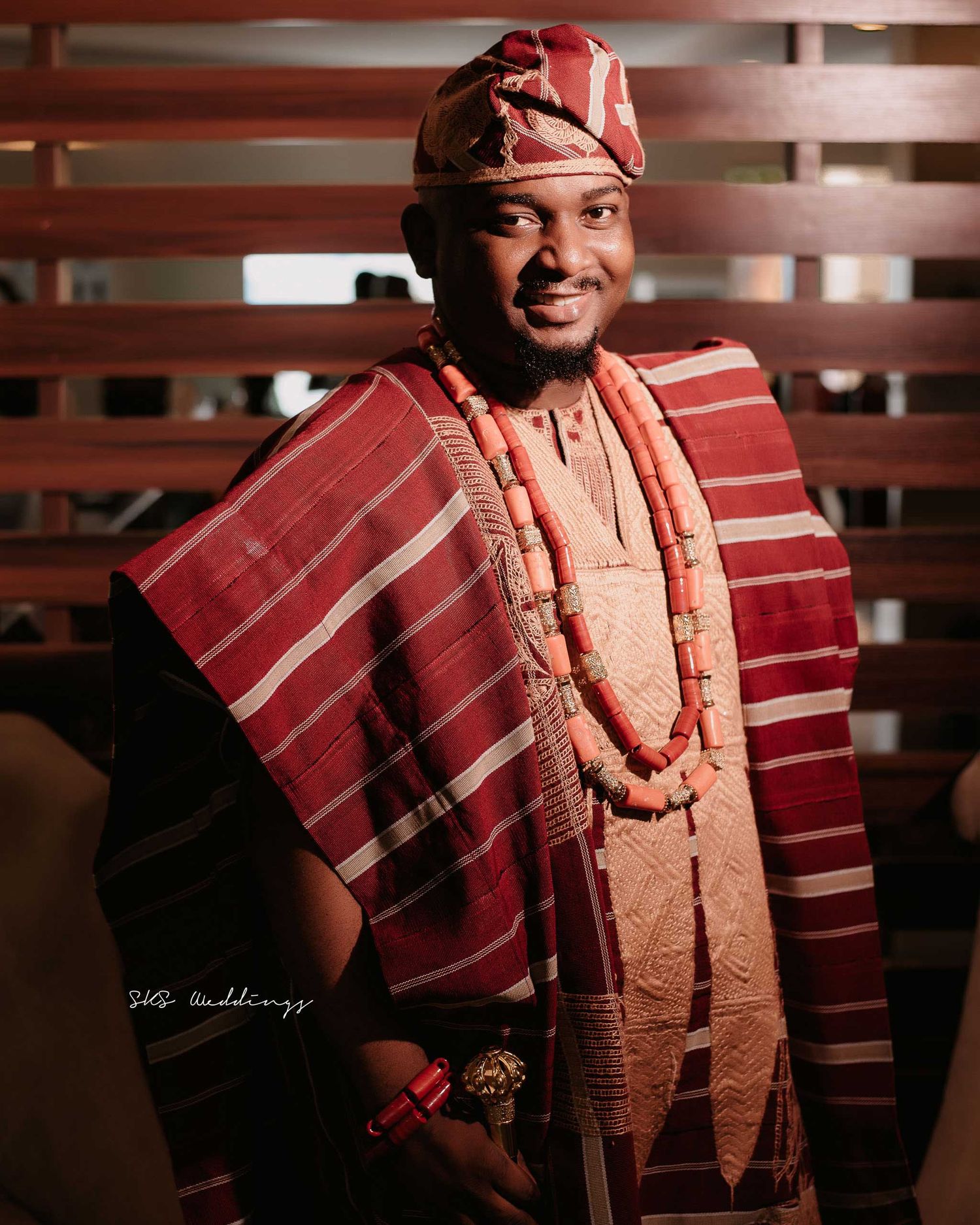 Traditional Nigerian groom wearing red and white Agbada with beaded coral necklaces and cap in formal portrait setting.
