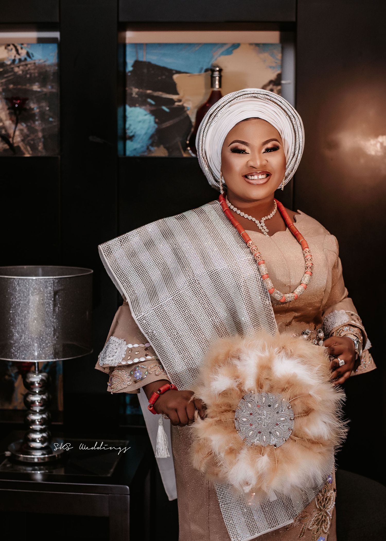 Traditional African wedding attire in peach tones with a matching gele headwrap and feather fan accessory.