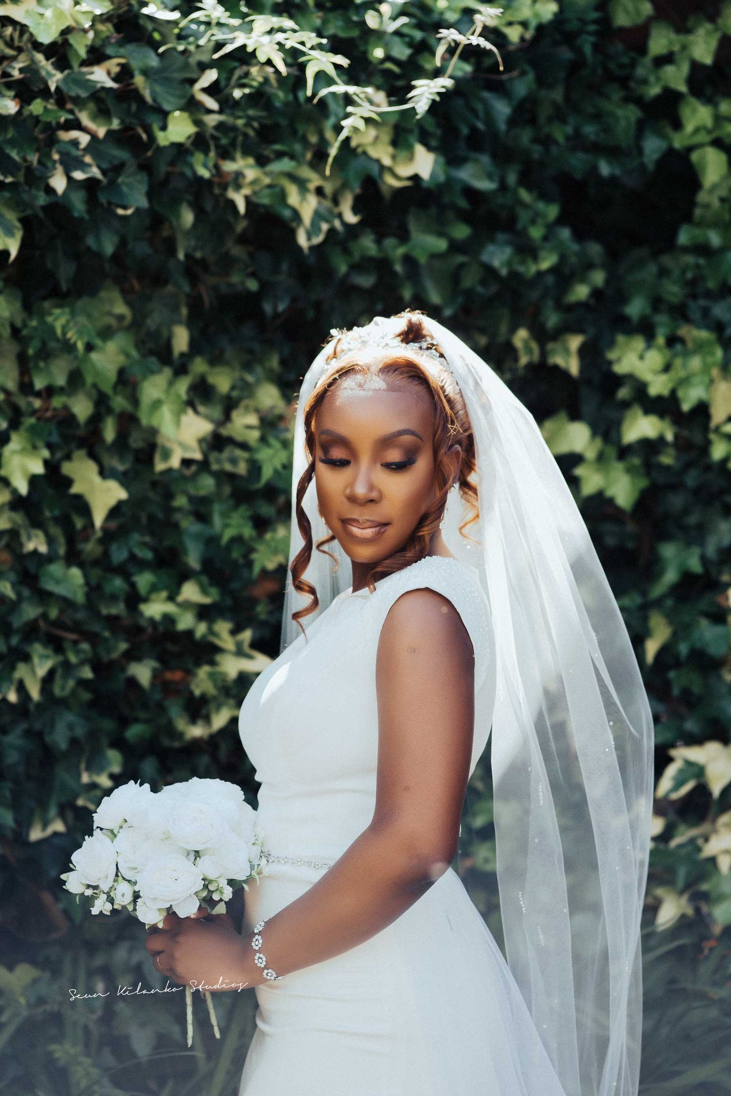 Bride in elegant white gown holds bouquet against garden backdrop.