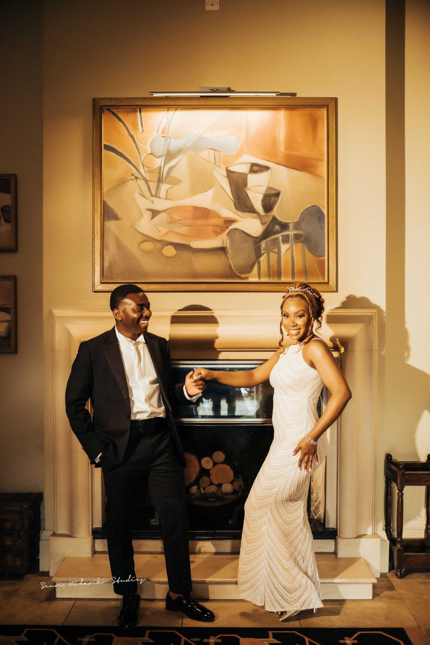Formal couple poses by fireplace with elegant artwork in warm-toned interior.