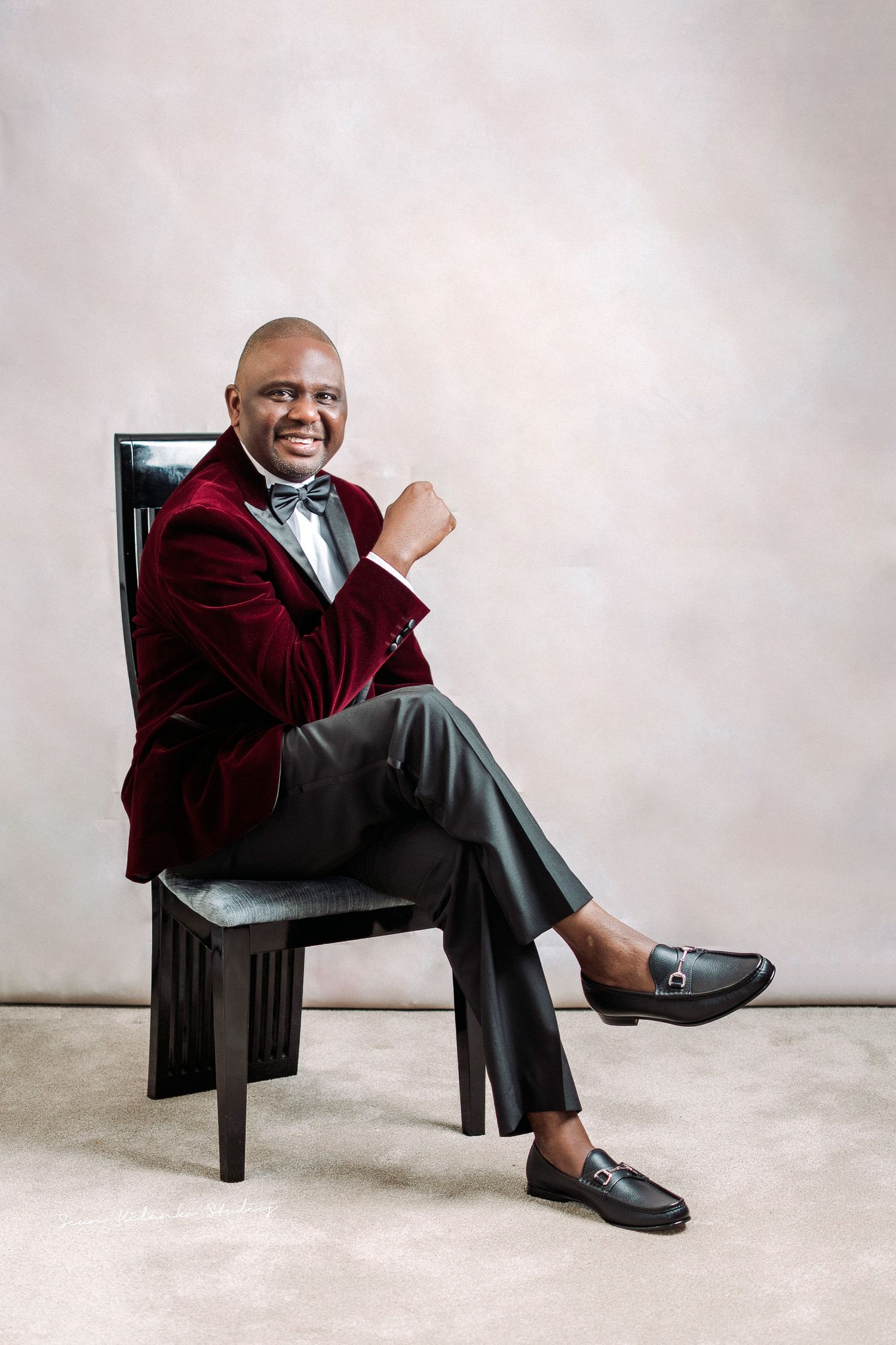 Elegant portrait in burgundy velvet blazer seated on black chair.