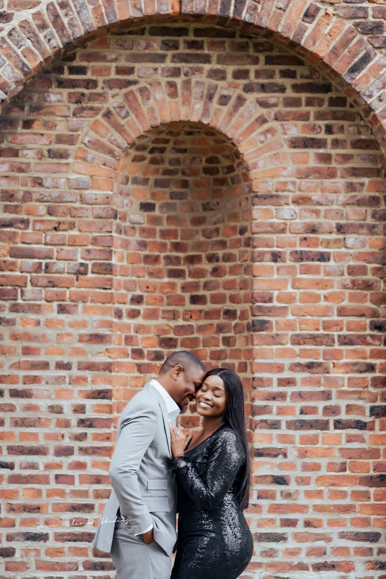 Romantic couple poses under brick archway during engagement shoot.