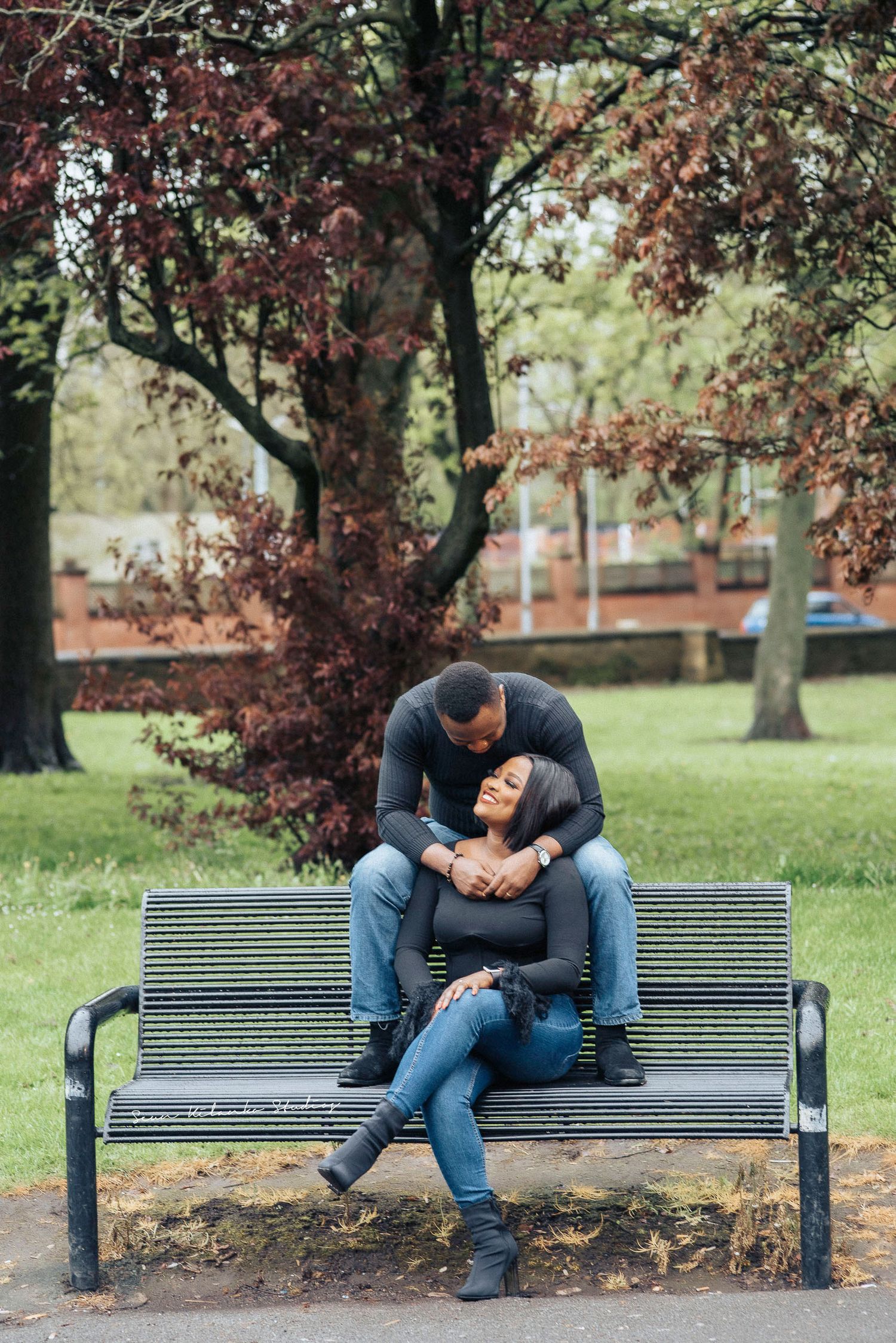 A couple shares an intimate moment while sitting on a park bench beneath autumn trees.