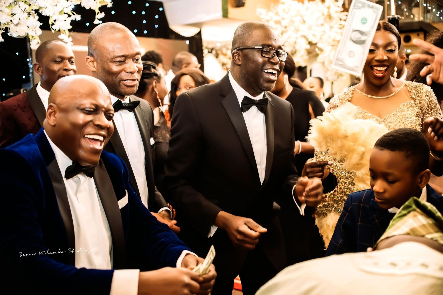 Guests in formal attire share a joyous moment at an elegant party or celebration with twinkling lights in the background.