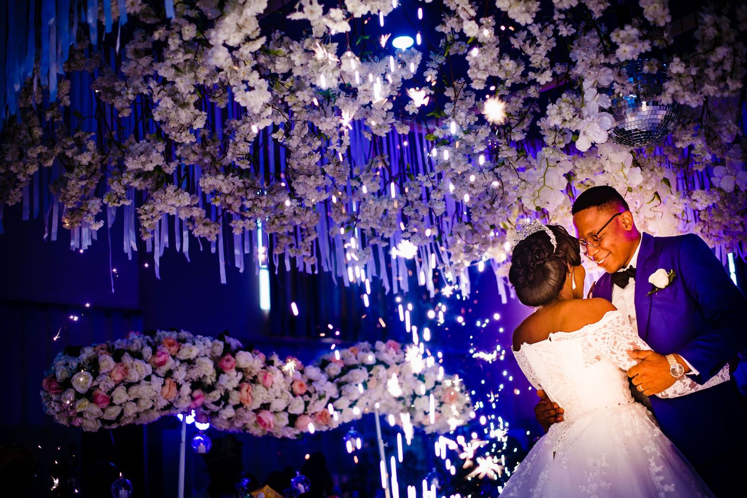 Elegant wedding couple dancing under suspended floral arrangements and hanging lights in a romantic blue-lit reception hall.